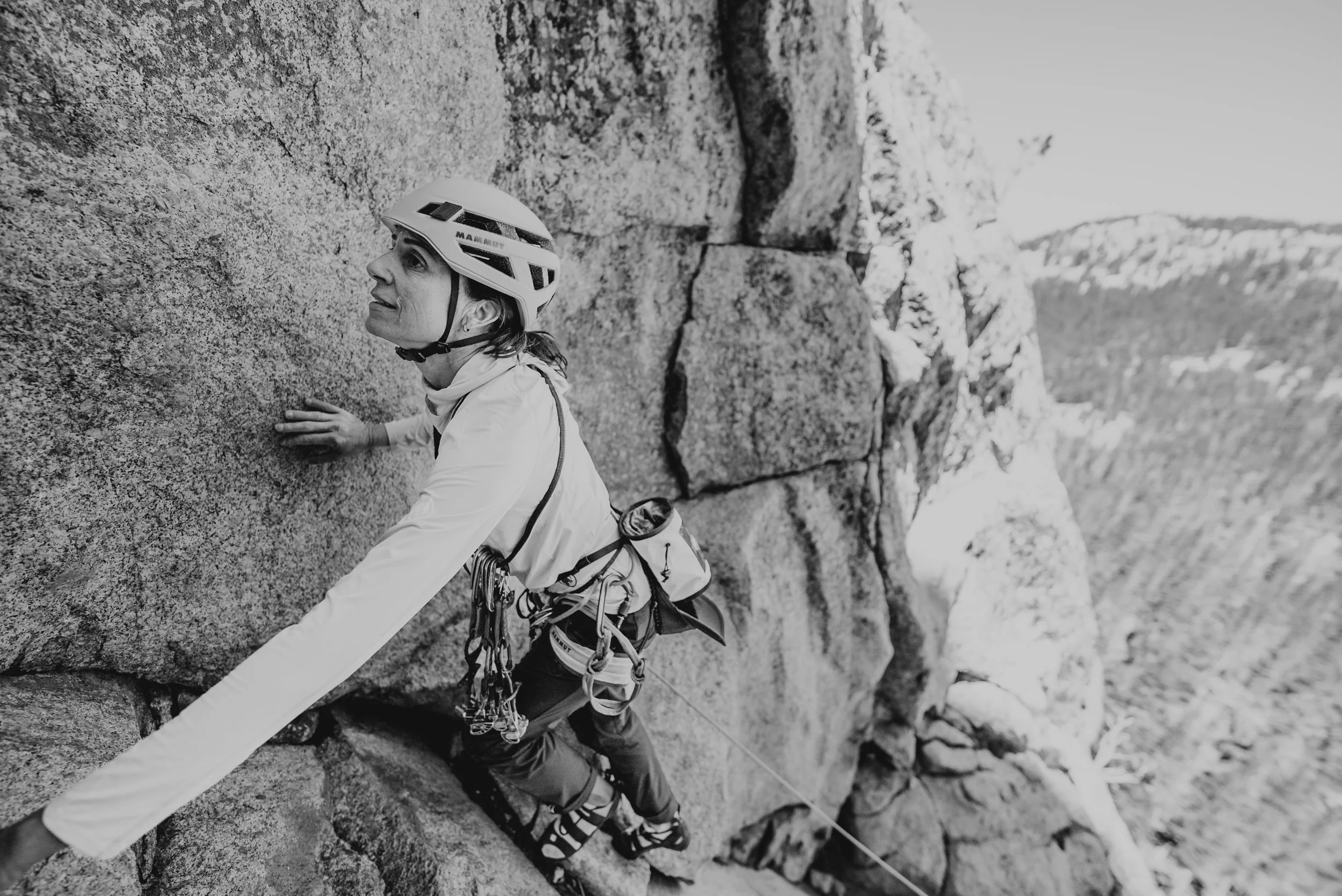 A woman rock climbing on a steep outdoor granite cliff, equipped with climbing gear and a helmet.