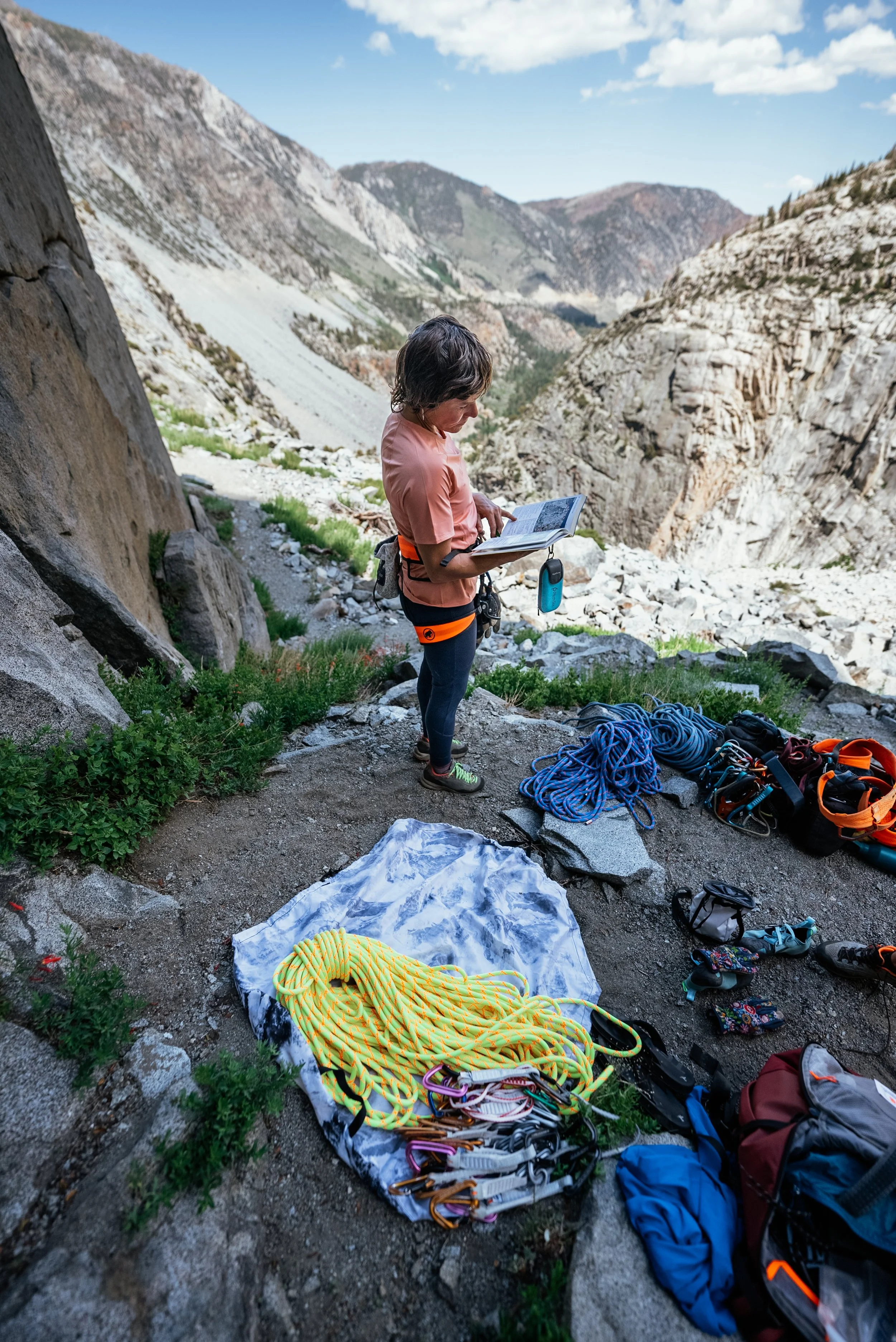 A woman stands on a mountain trail reading a guidebook, surrounded by climbing gear including ropes, harnesses, and carabiners, with a canyon and mountains in the background.