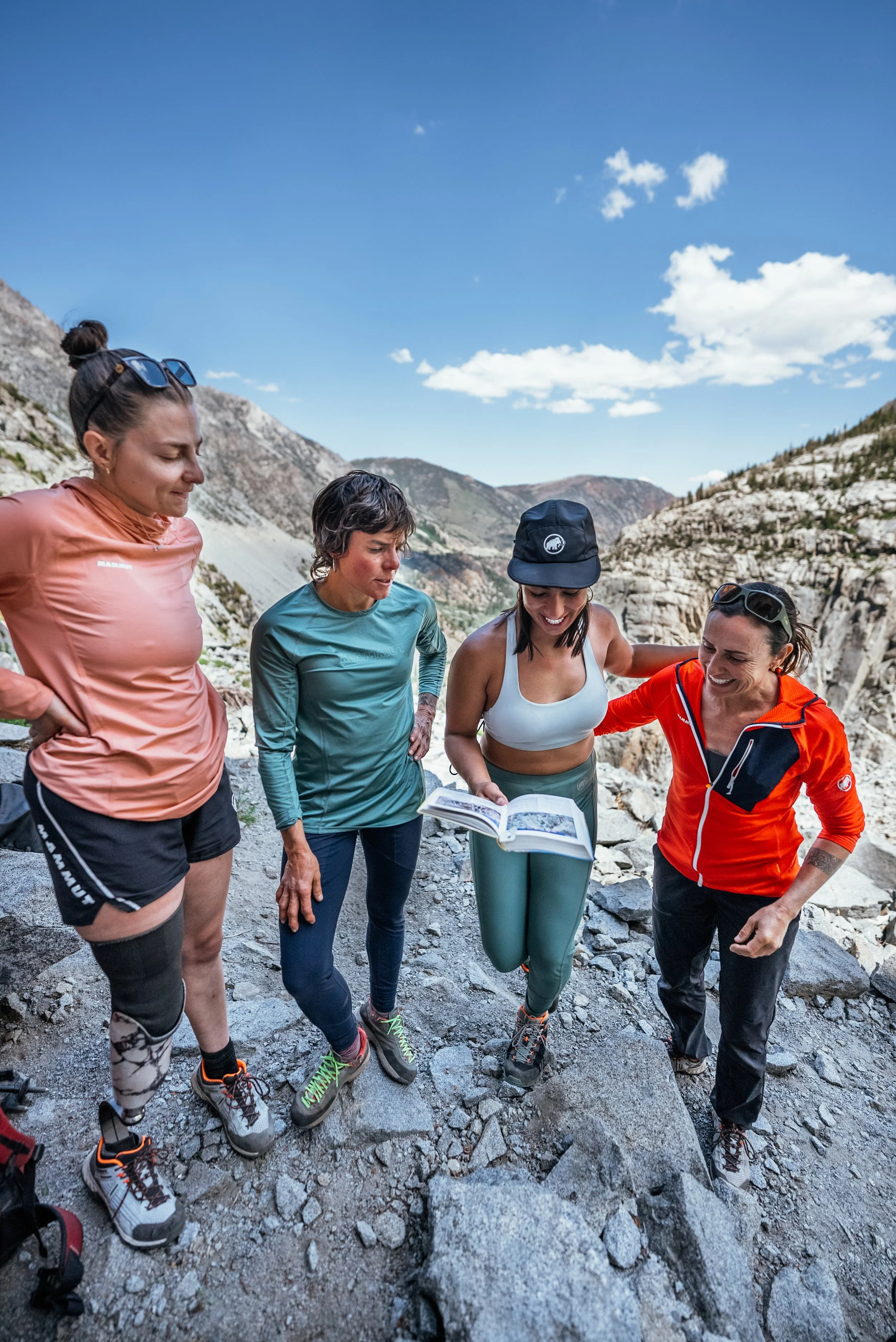 Four women on a rocky mountain trail looking at a map, enjoying a hike in rugged terrain under a blue sky with clouds.