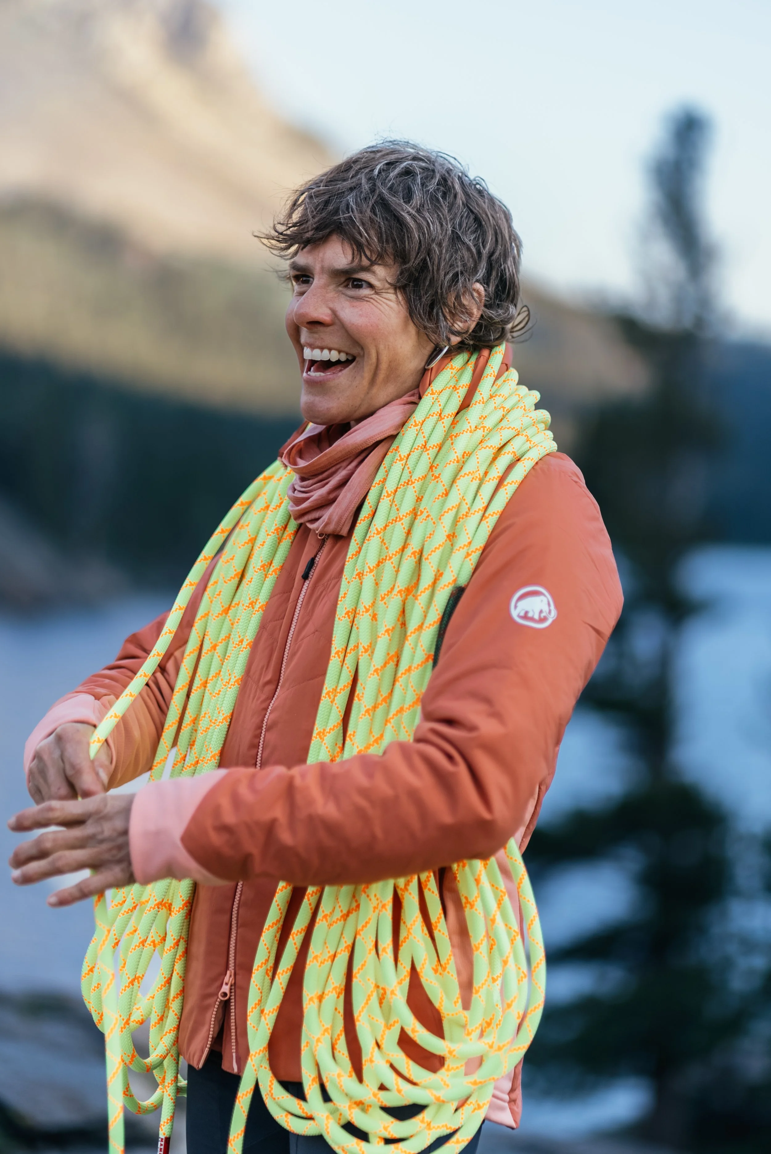 A woman with short curly hair wearing an orange jacket and a pink scarf, smiling and holding yellow and orange patterned ropes outdoors near water and trees.