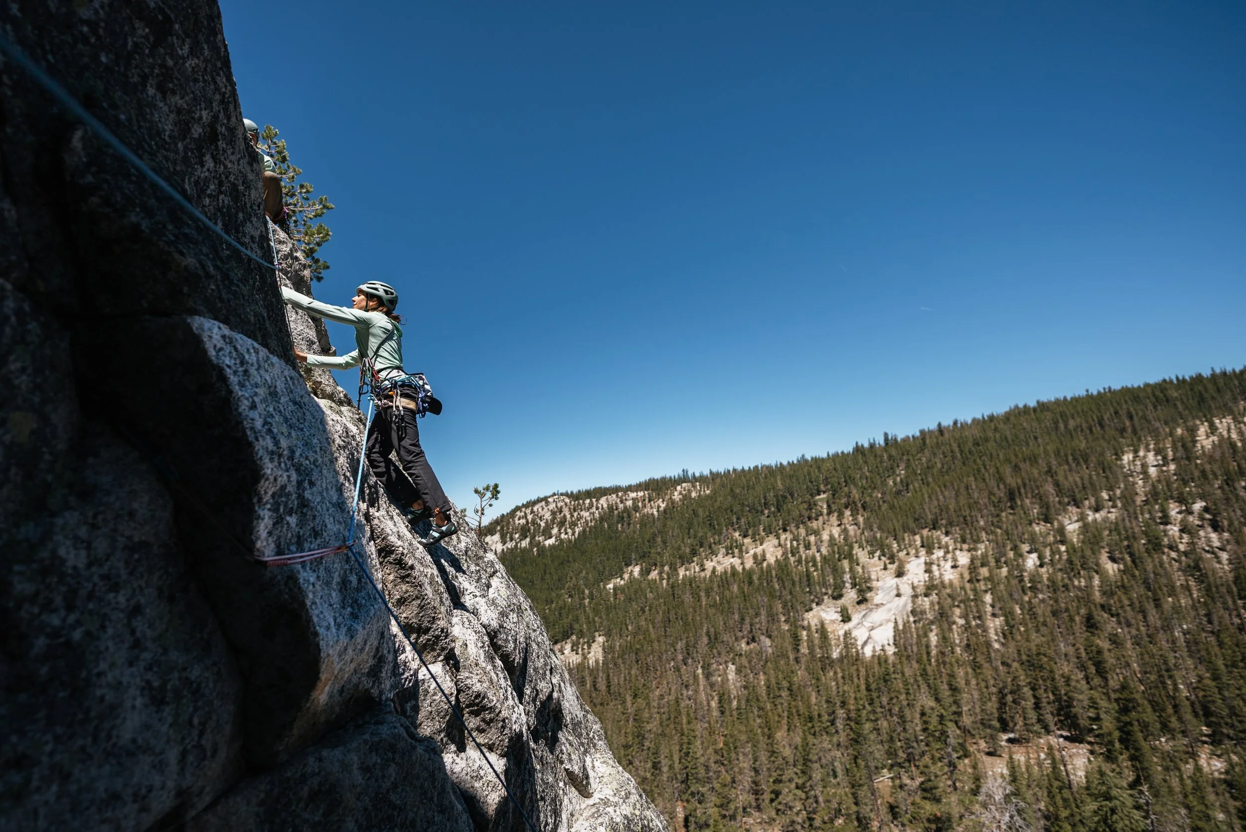 A person rock climbing on a rugged mountain face, wearing a helmet and climbing gear, with a forested mountain in the background under a clear blue sky.