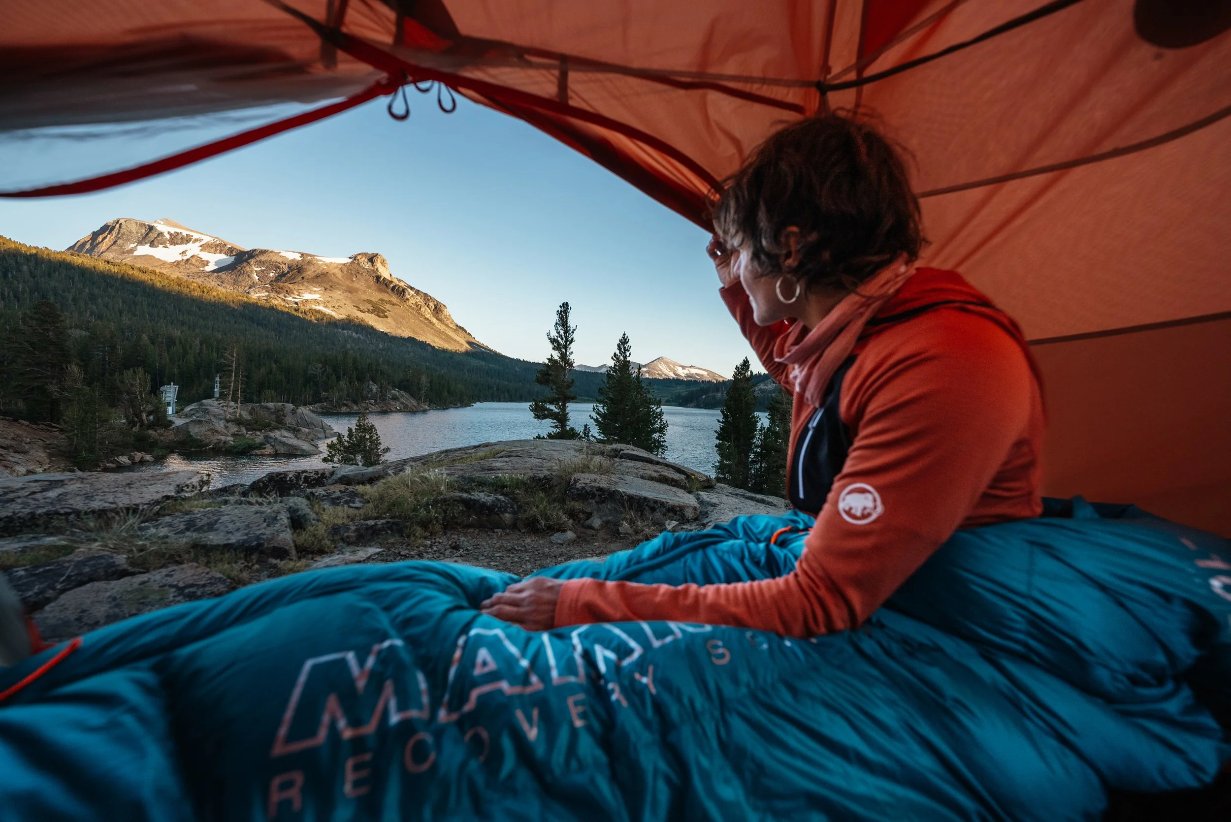 Person inside a tent, looking out at a mountain lake scene with pine trees and snow-capped peaks.