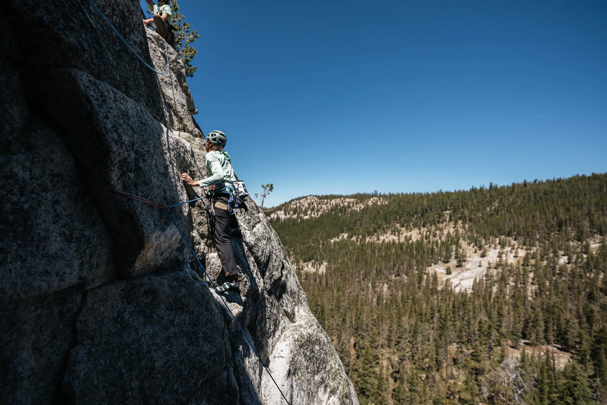 A person wearing a helmet and climbing gear is on a rock face, securing a safety rope during outdoor rock climbing in a mountainous area with a forest below under a clear blue sky.