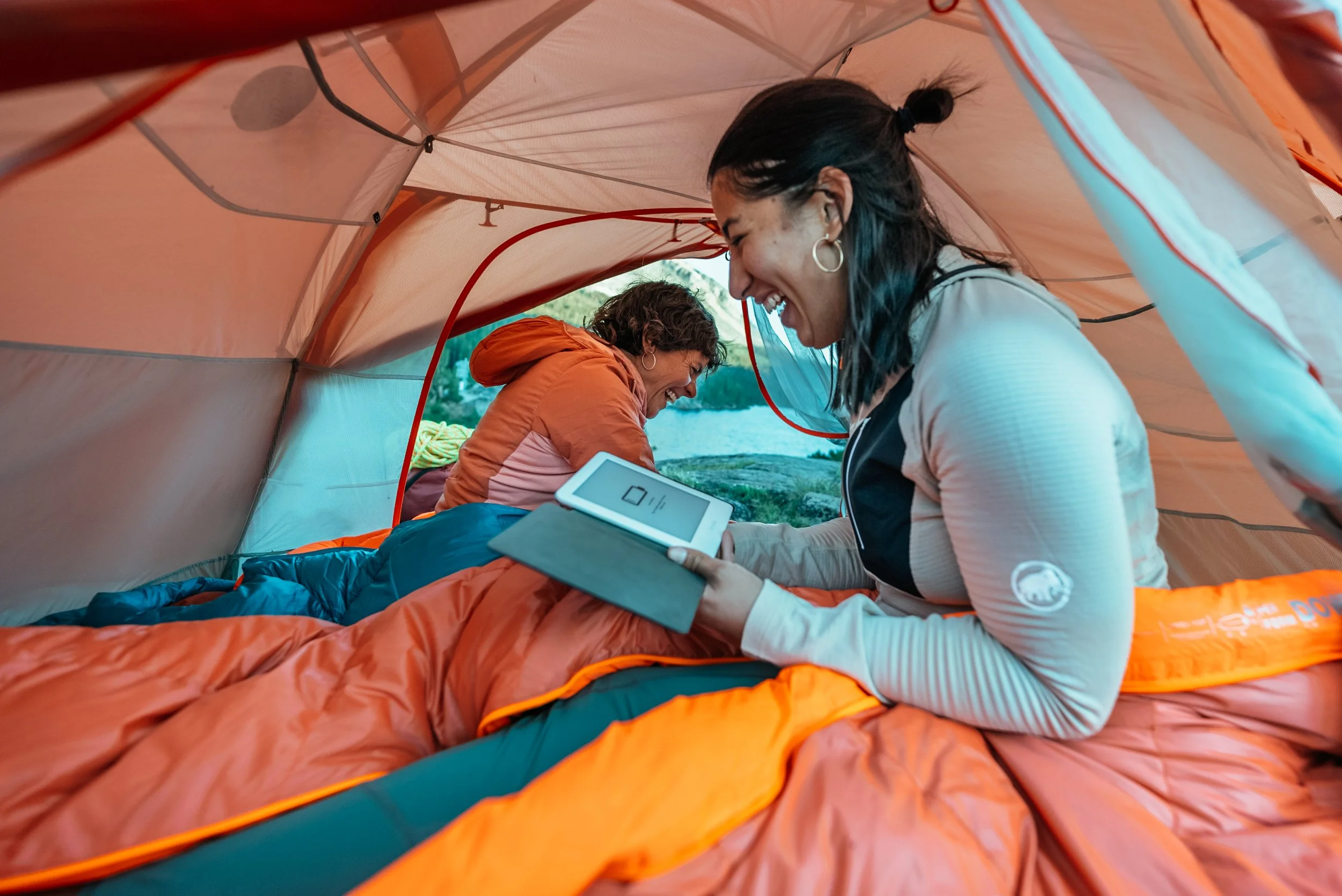 Two women inside a camping tent smiling and sharing a happy moment, with one using a tablet. The tent is orange and they are outdoors near a body of water.