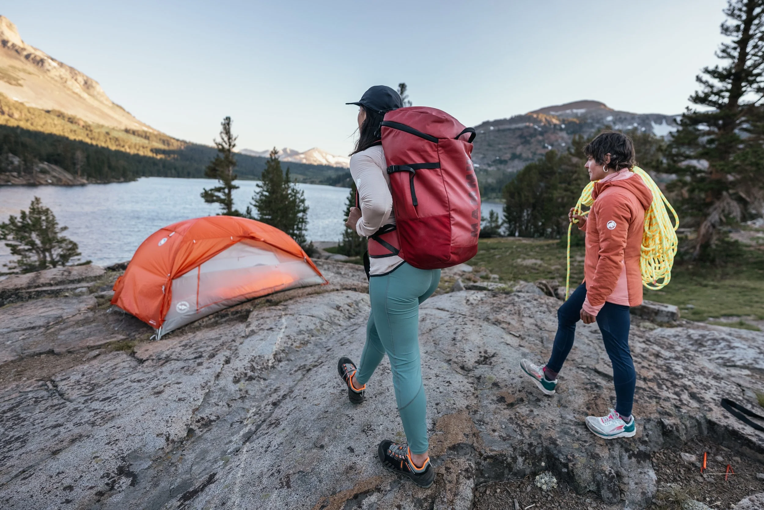 Two women with backpacks hiking near a lake with a tent on a rocky terrain, surrounded by trees and mountains during sunset.