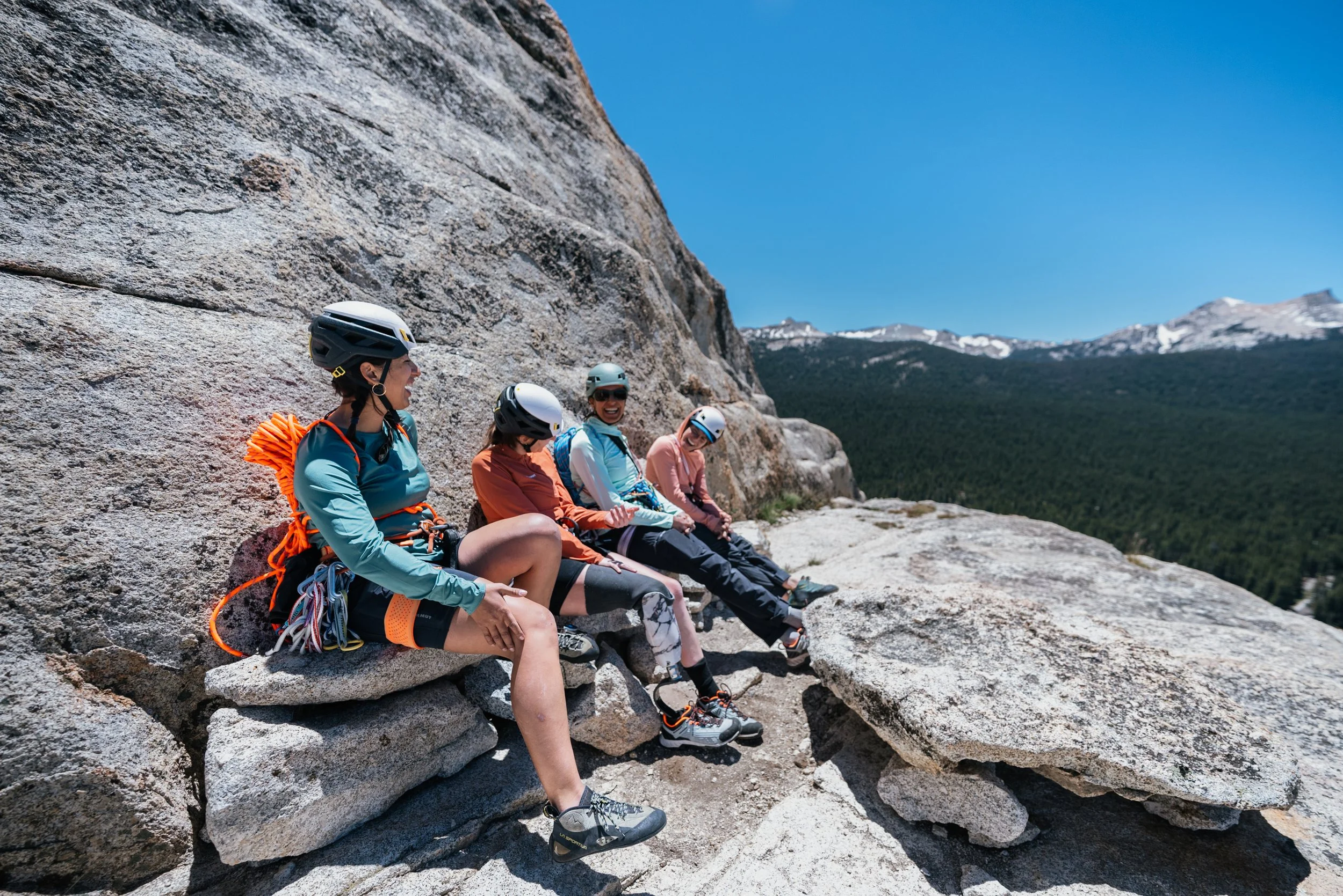 Four women wearing helmets and outdoor gear sitting on a large rock ledge overlooking a mountain landscape with blue sky and snowy peaks.