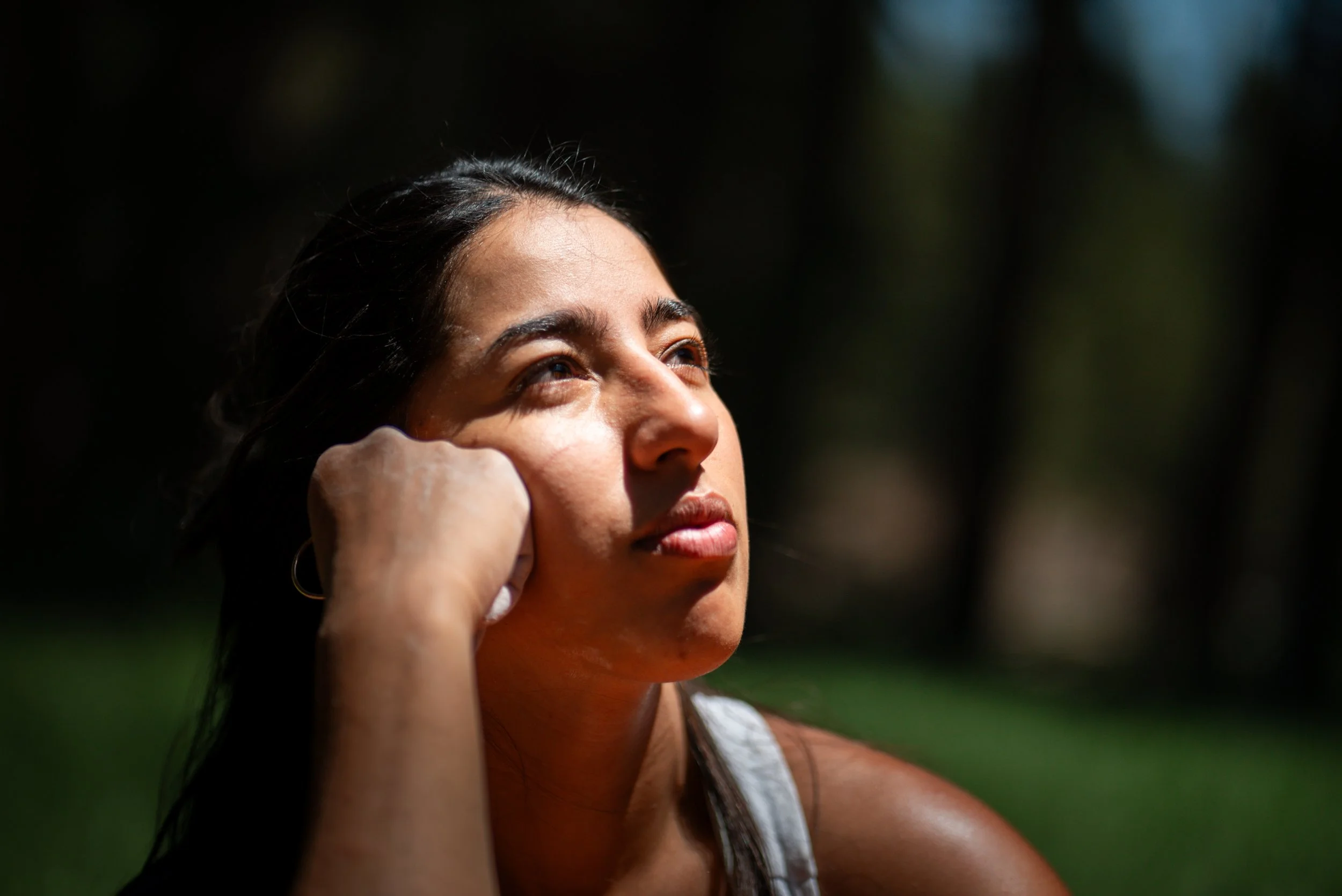 Close-up of a woman with dark hair looking upward in sunlight, with a blurred green background.