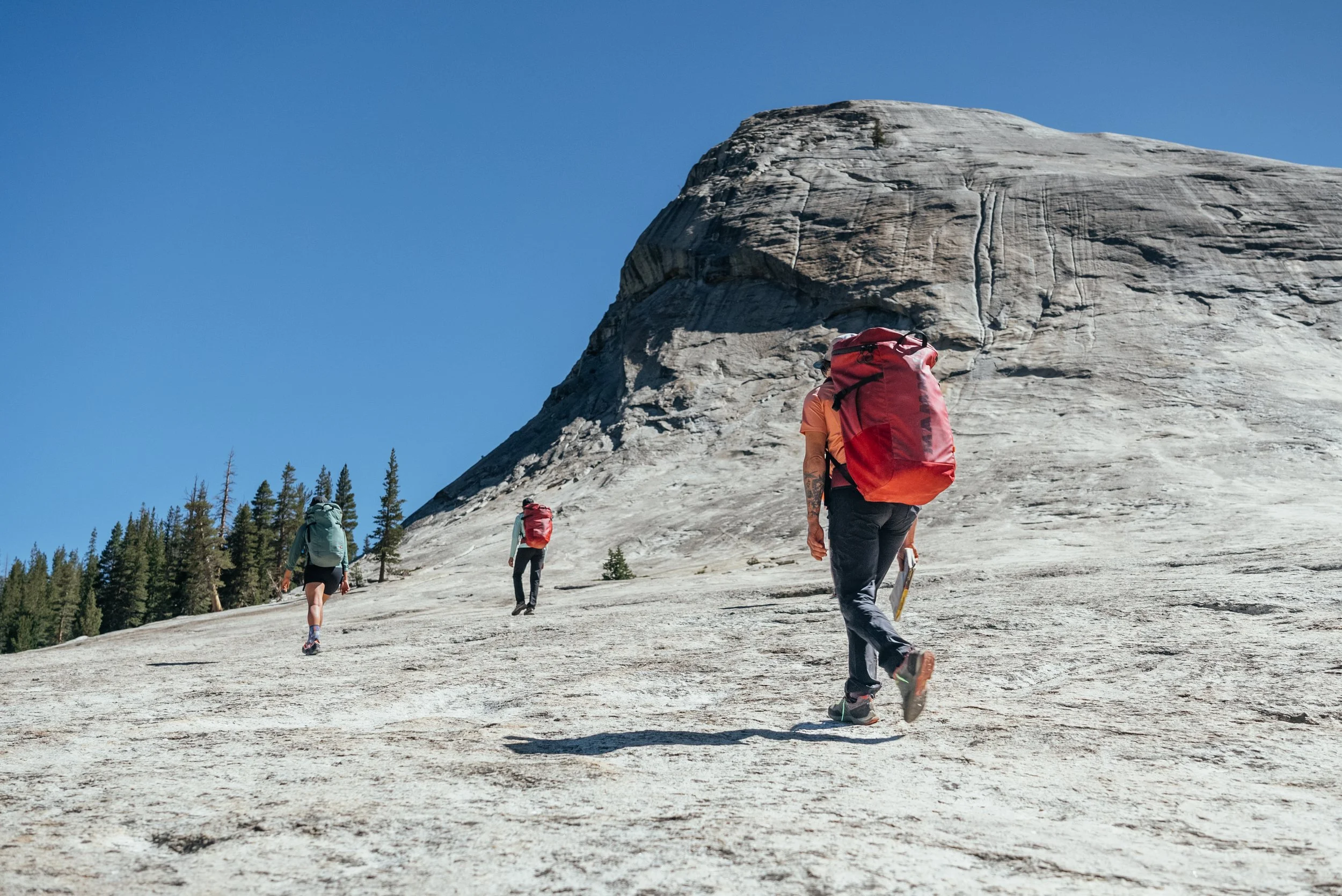 Three hikers with backpacks walking up a rocky mountain slope with a large rock formation in the background and a clear blue sky.