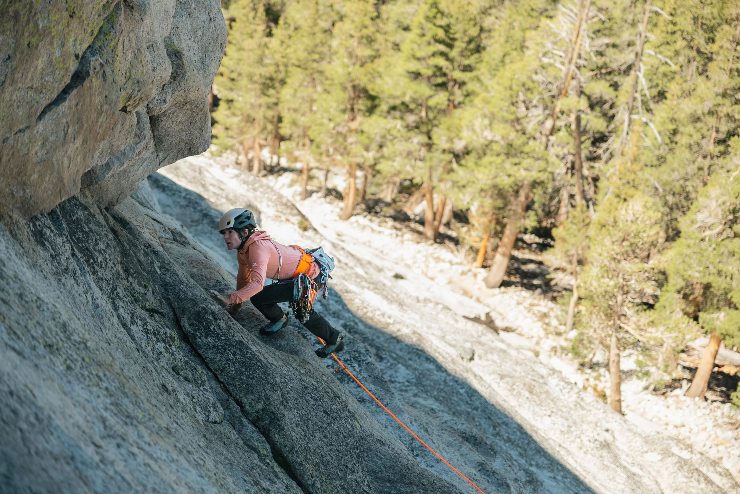 A woman rock climbing on a steep granite face in a forested area, wearing a helmet and safety gear.