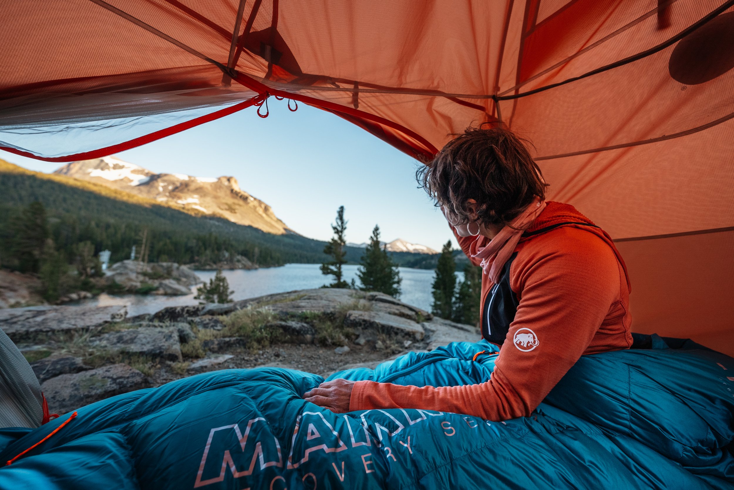A person inside a tent overlooking a mountain lake, wearing an orange jacket and sitting on a blue sleeping bag.
