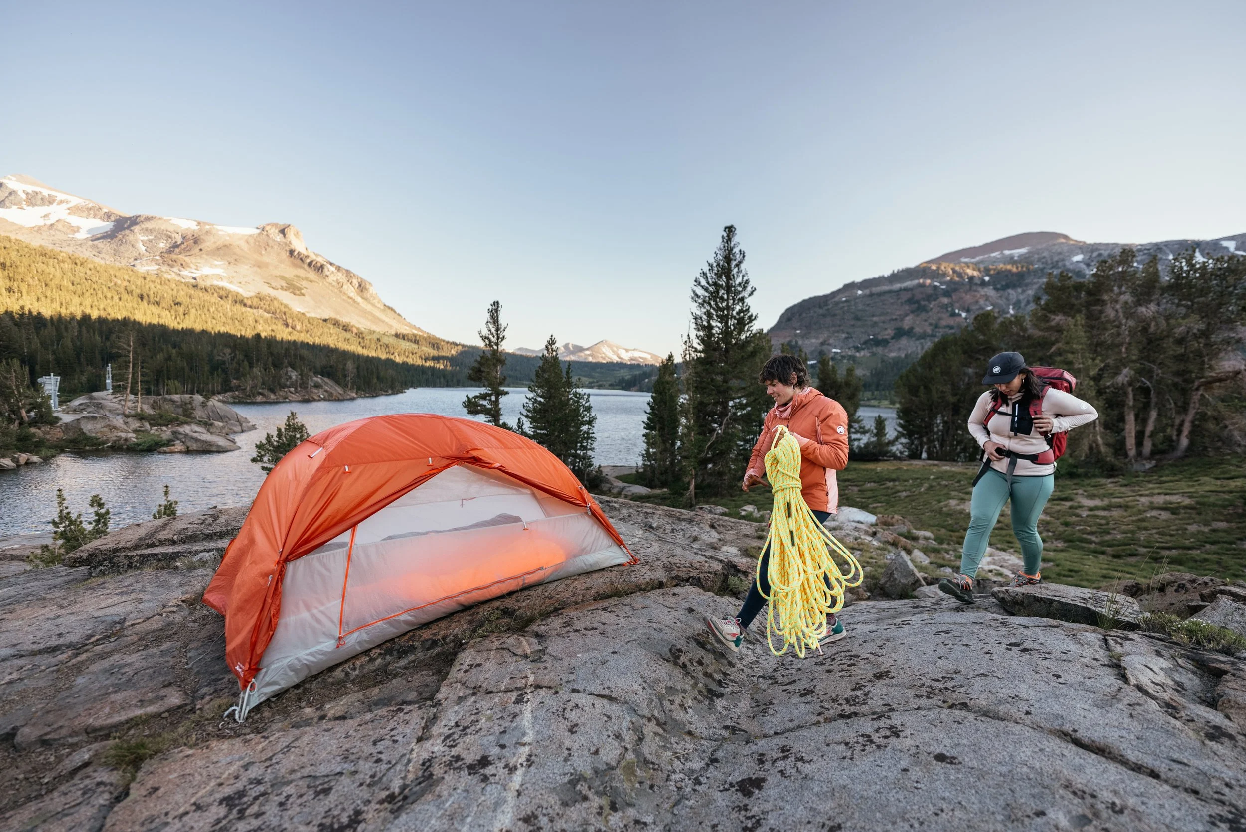 Two people preparing camping gear near a lake in a mountainous forest setting, with a tent on the rocky ground.
