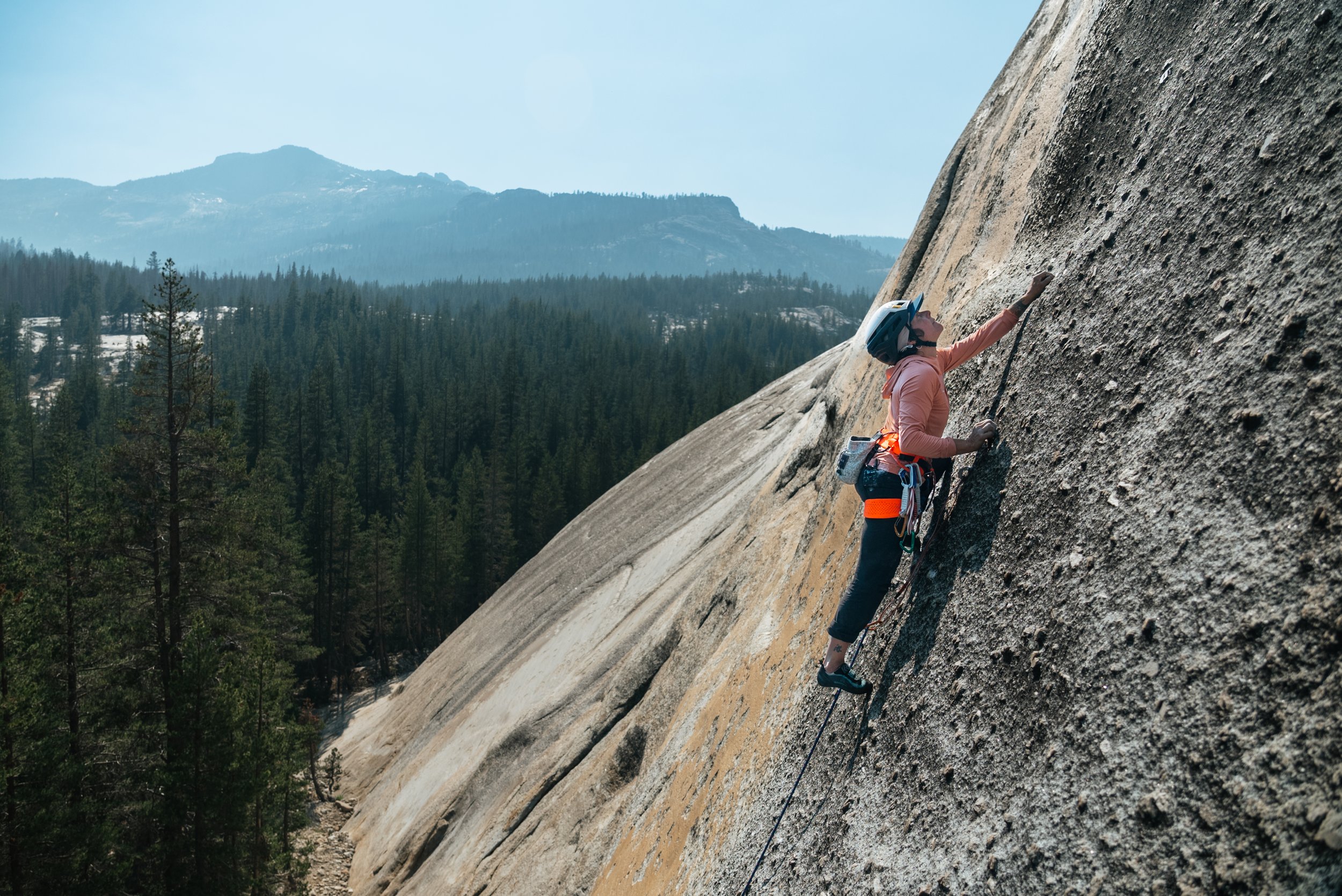 A woman climbing a steep rock face outdoors, wearing a helmet and climbing gear with a harness.