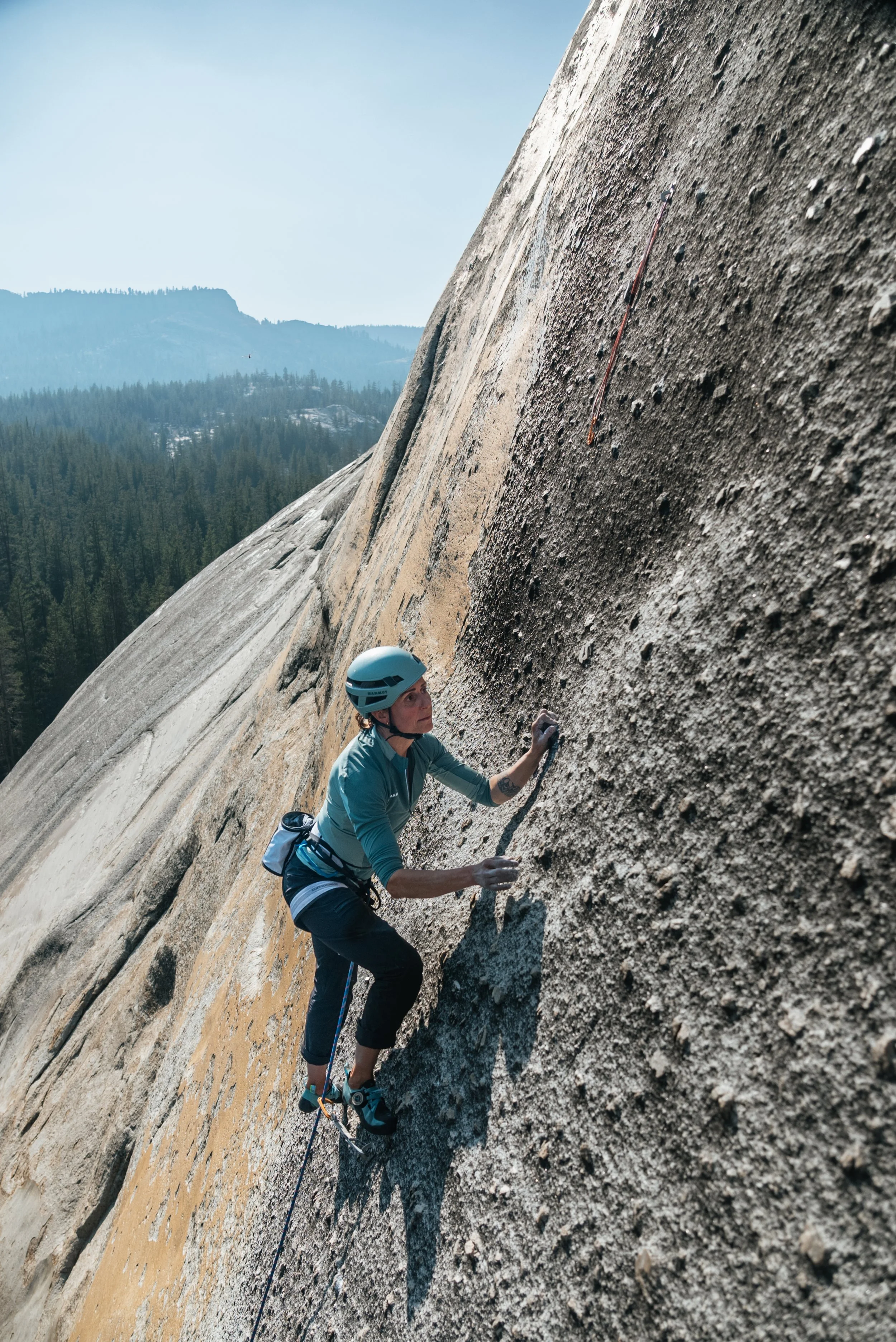 A woman rock climbing on a steep granite wall in an outdoor setting with trees and mountains in the background.