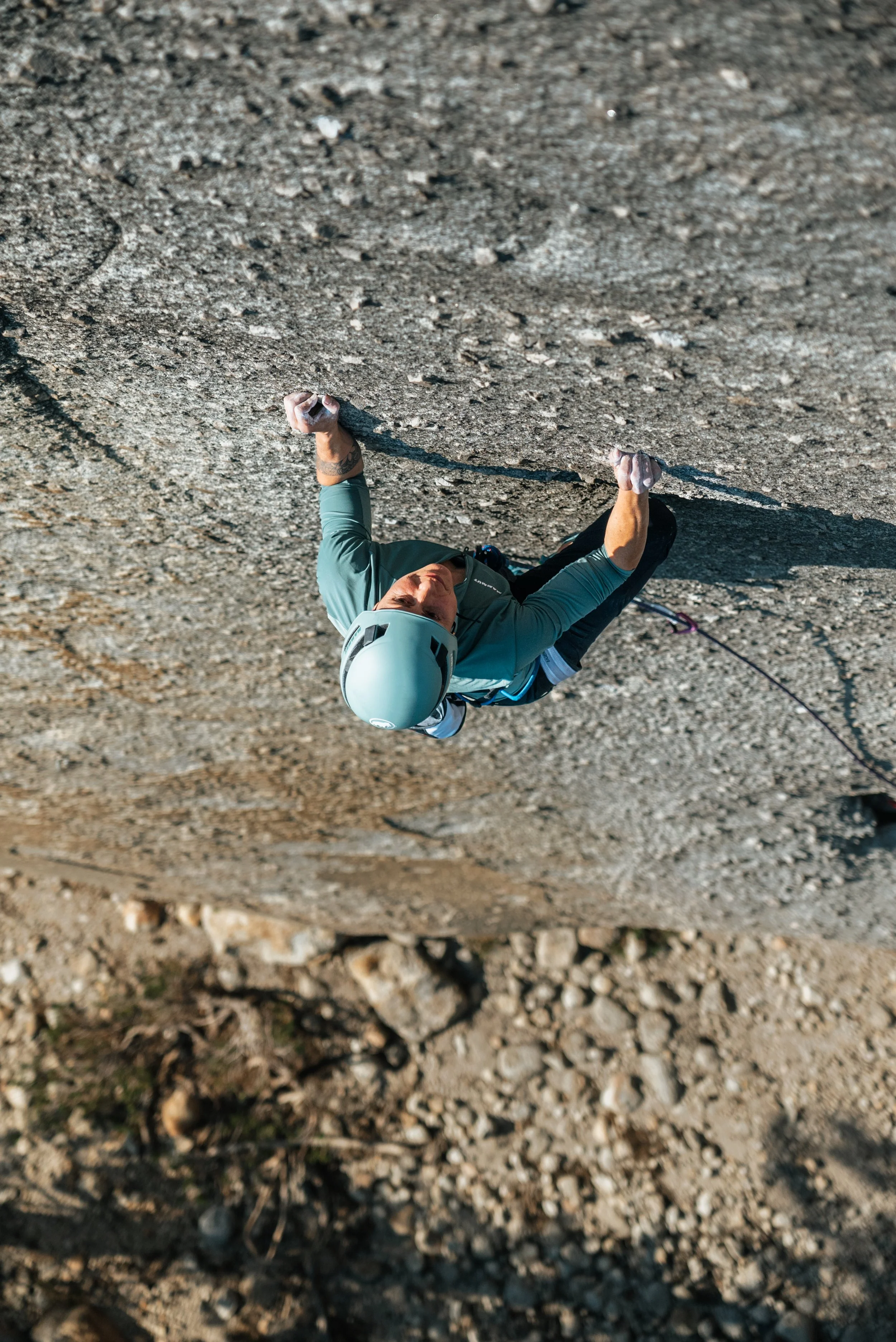 Climber wearing a helmet and teal jacket gripping a rock face during a climb on a rocky outdoor surface.