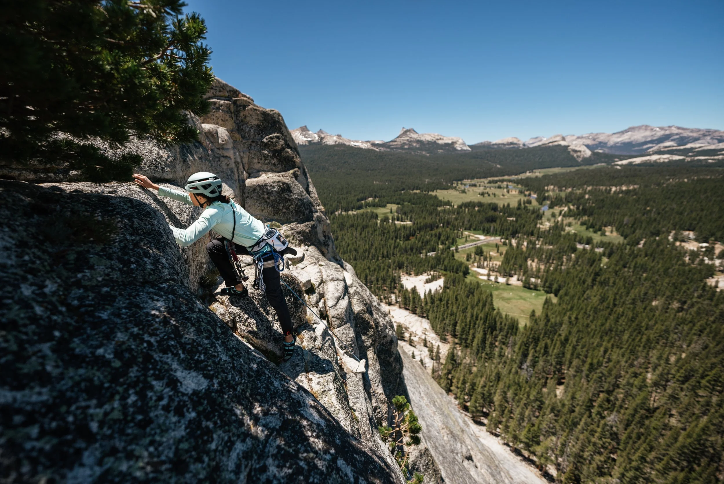 A woman rock climbing on a steep mountain face with a forested valley and mountains in the background.