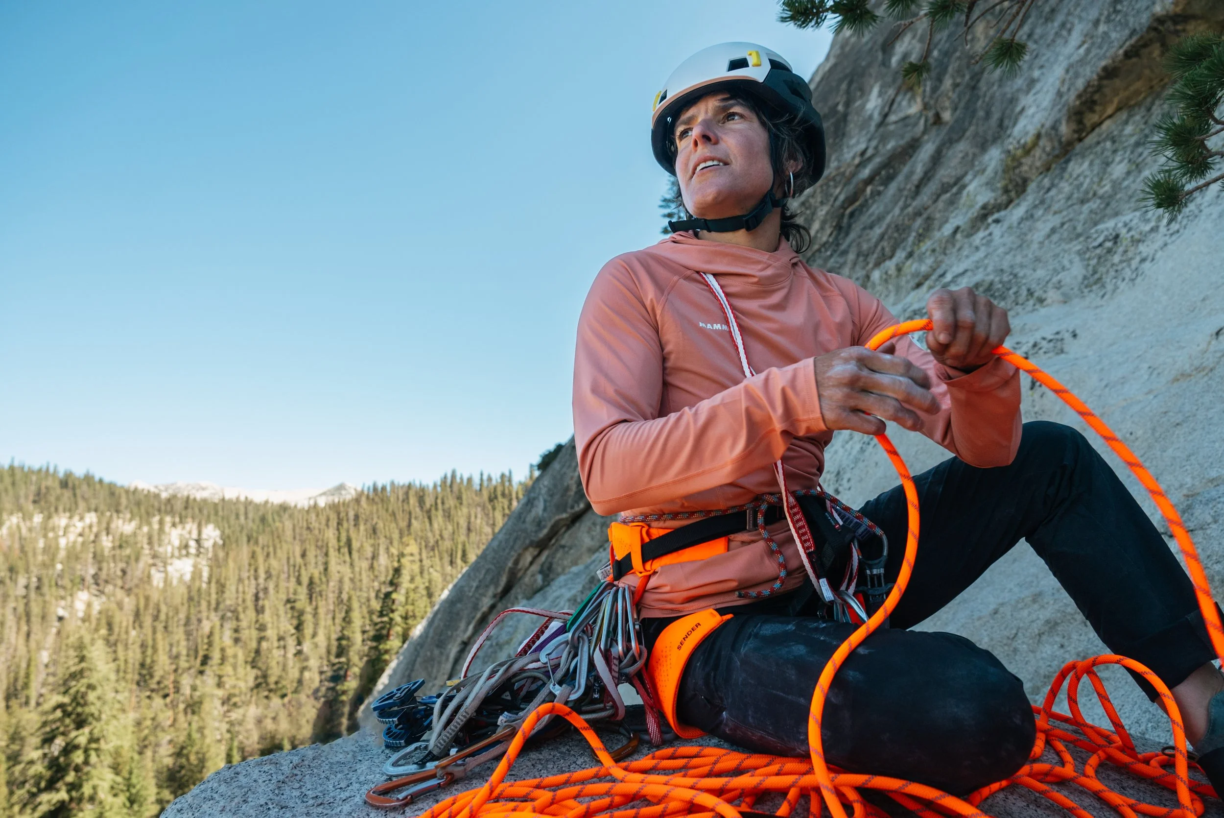 A woman climber sitting on a rock face with climbing gear, holding an orange climbing rope, wearing a white helmet, pinkish jacket, and black pants, with a forested landscape in the background.