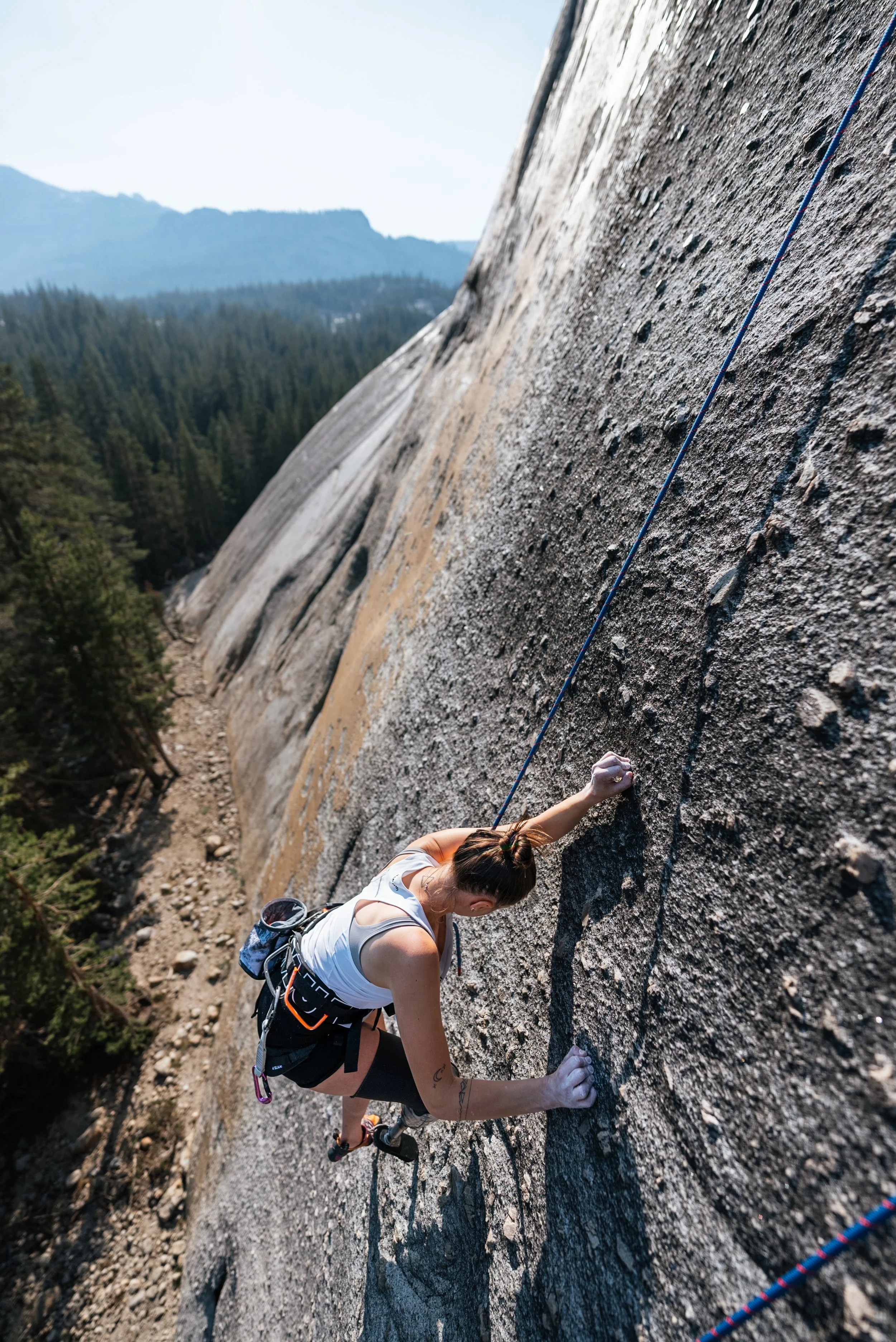 A woman rock climbing on a steep granite wall in a mountainous forested area during daytime.