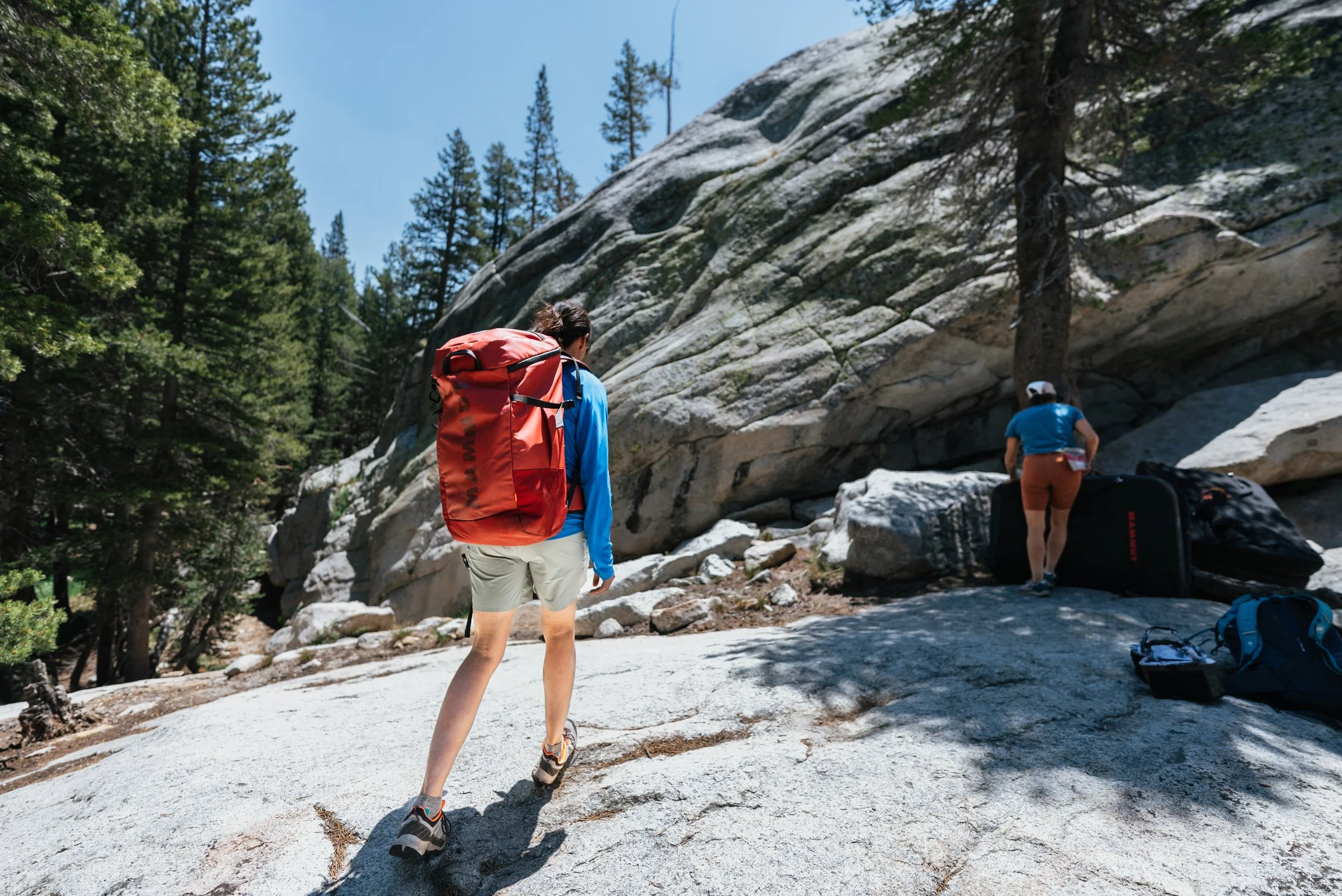 Two women hiking in a mountain forest, with one woman carrying a large red backpack and another near a large black bag, surrounded by tall trees and rocky terrain.