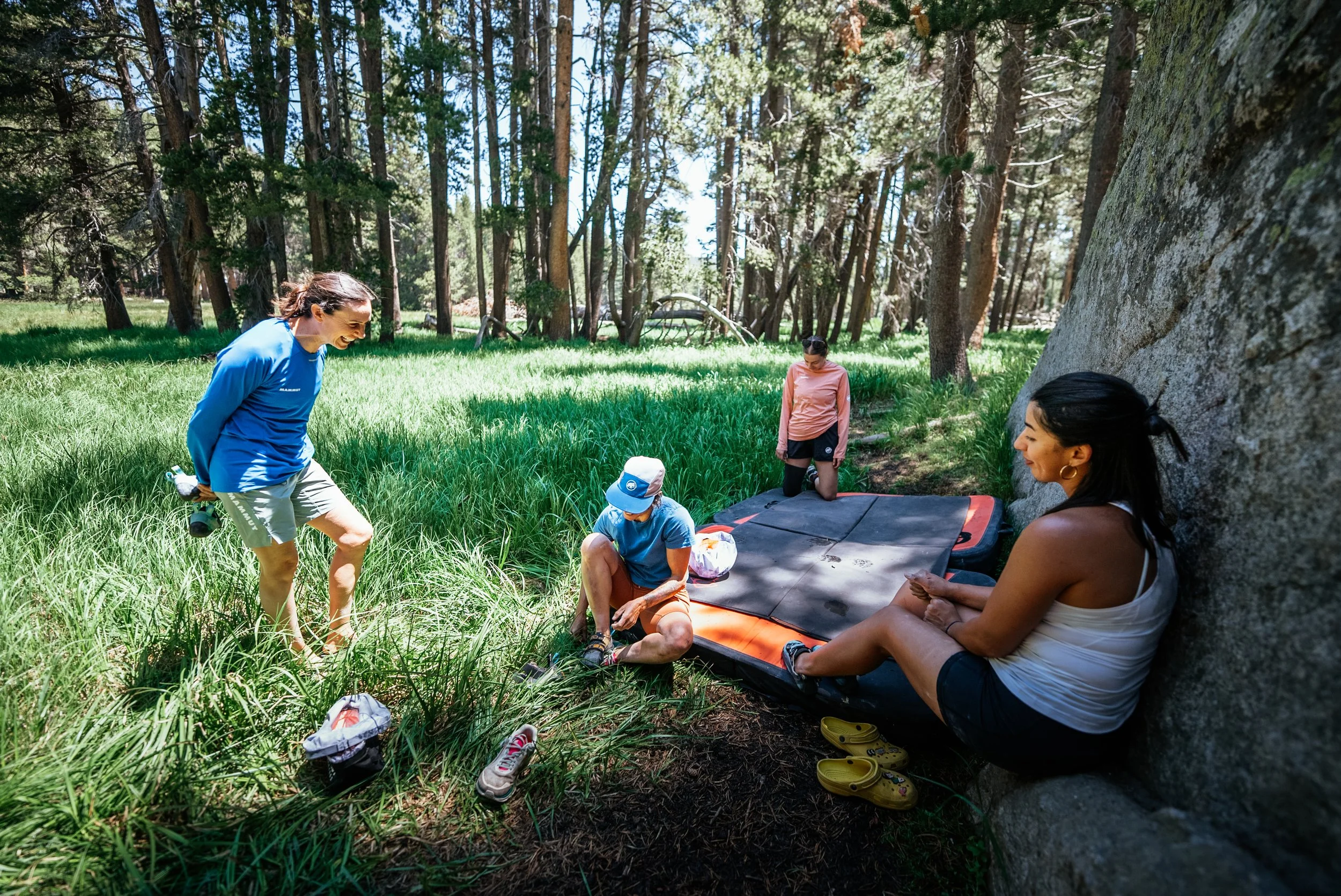 Group of friends in a forest clearing, some sitting on a boulders and a mat, others standing, enjoying a sunny day in the woods with tall trees and lush grass.