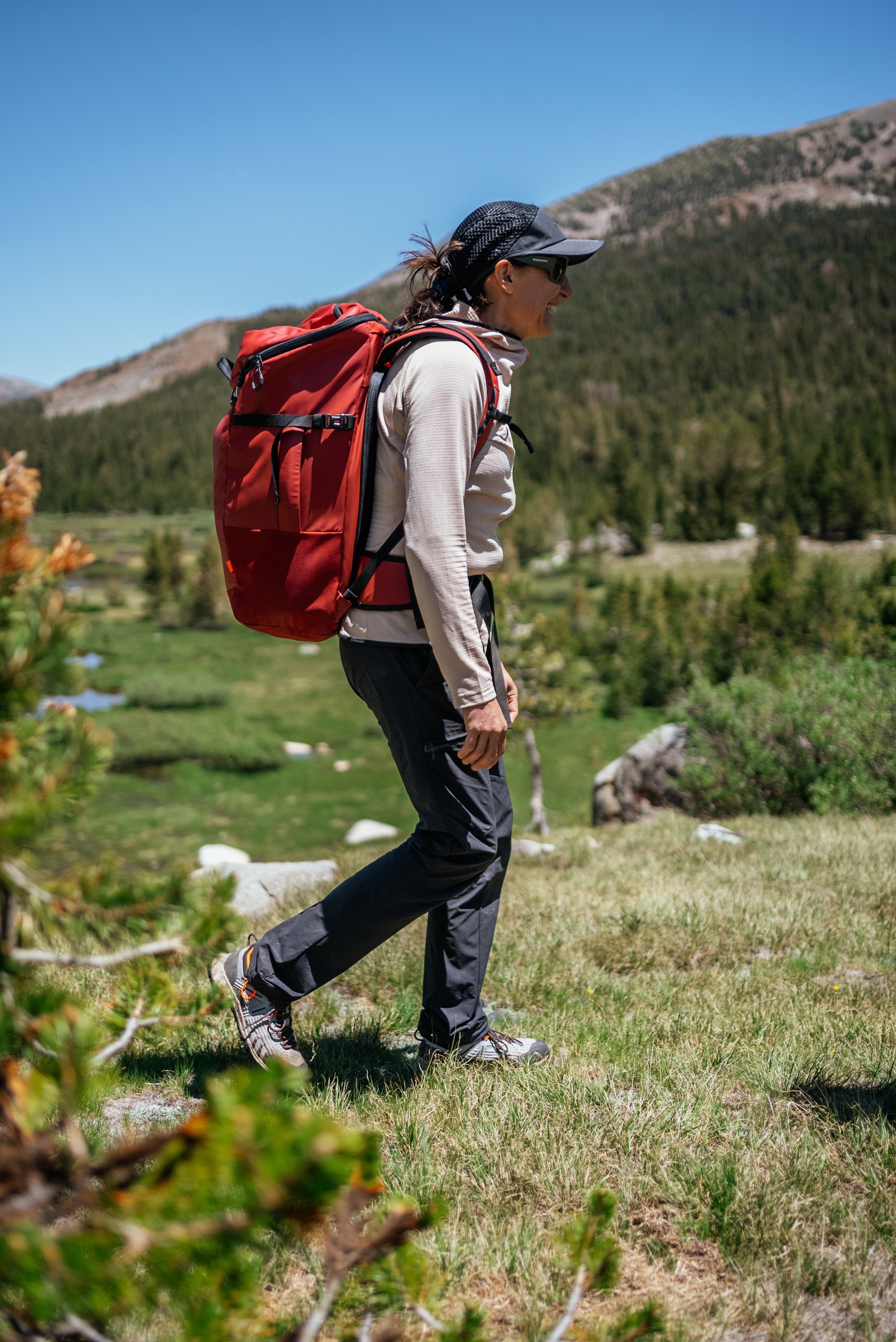 A woman hiking in a mountainous area with a large red backpack, wearing a hat, sunglasses, and outdoor clothing, on a clear, sunny day.