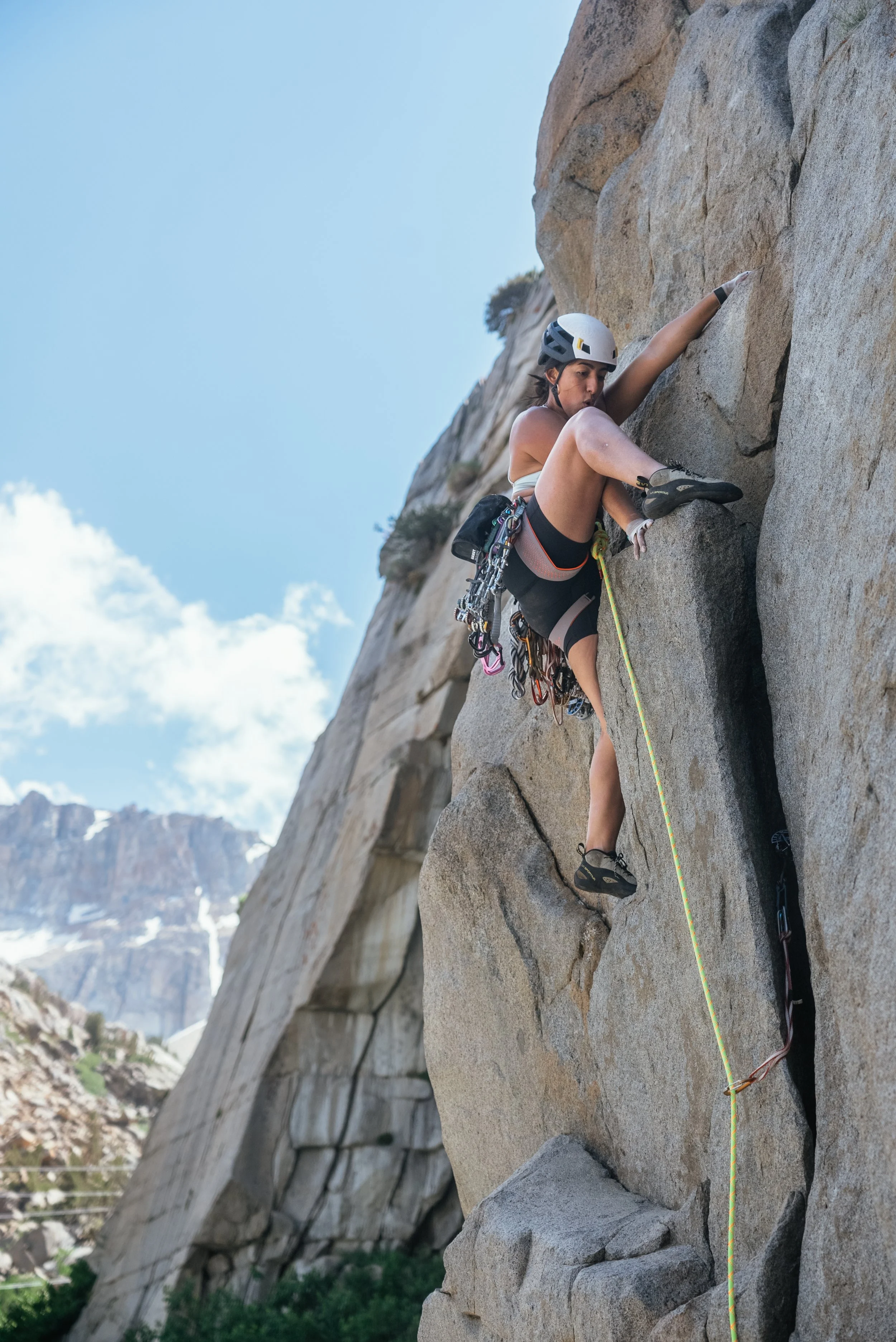 A woman climbing a rock face outdoors, wearing a helmet and harness, with climbing gear attached to her waist.