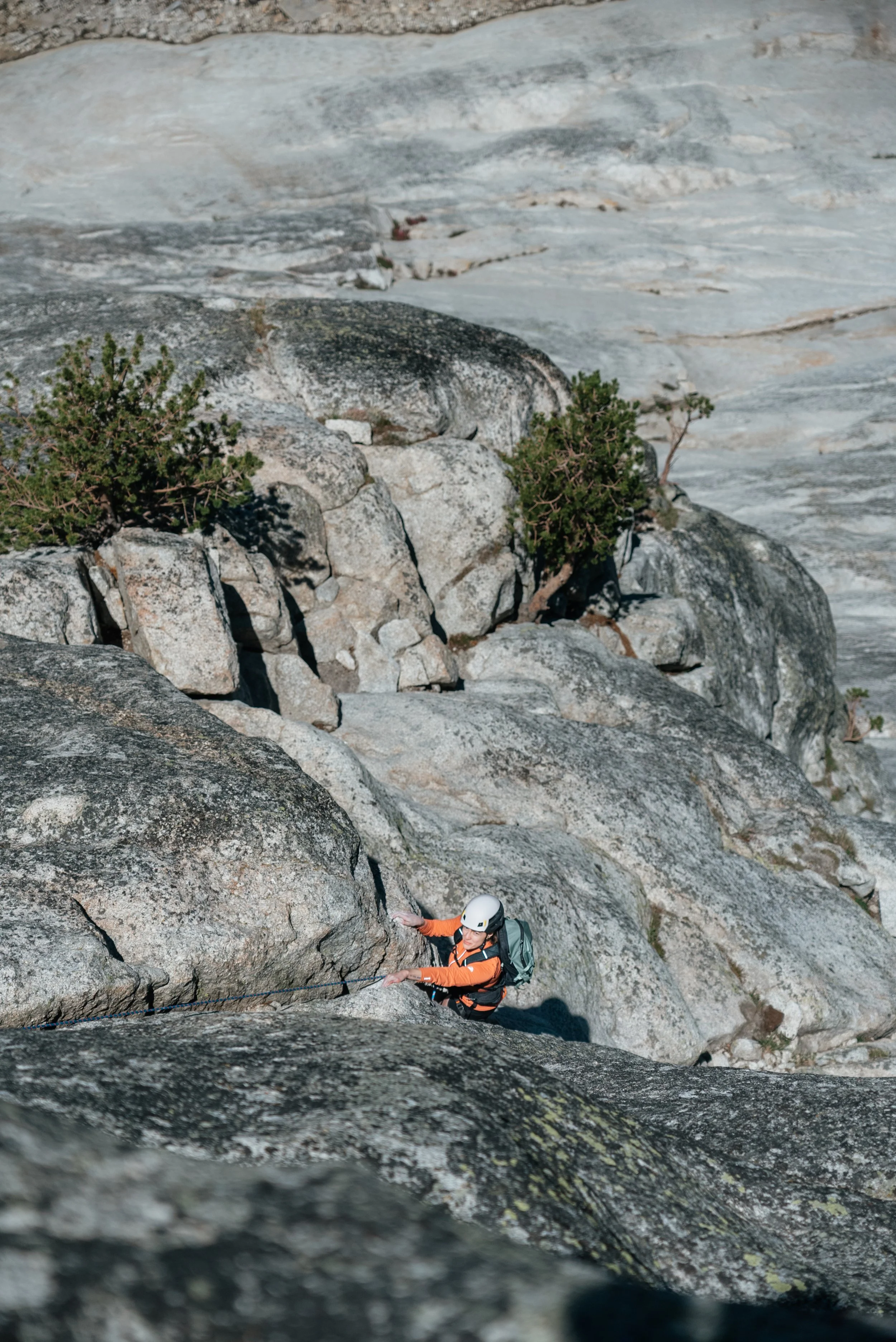 A person wearing a helmet, orange jacket, and backpack rock climbing on a gray granite cliff with shrubs and trees nearby.