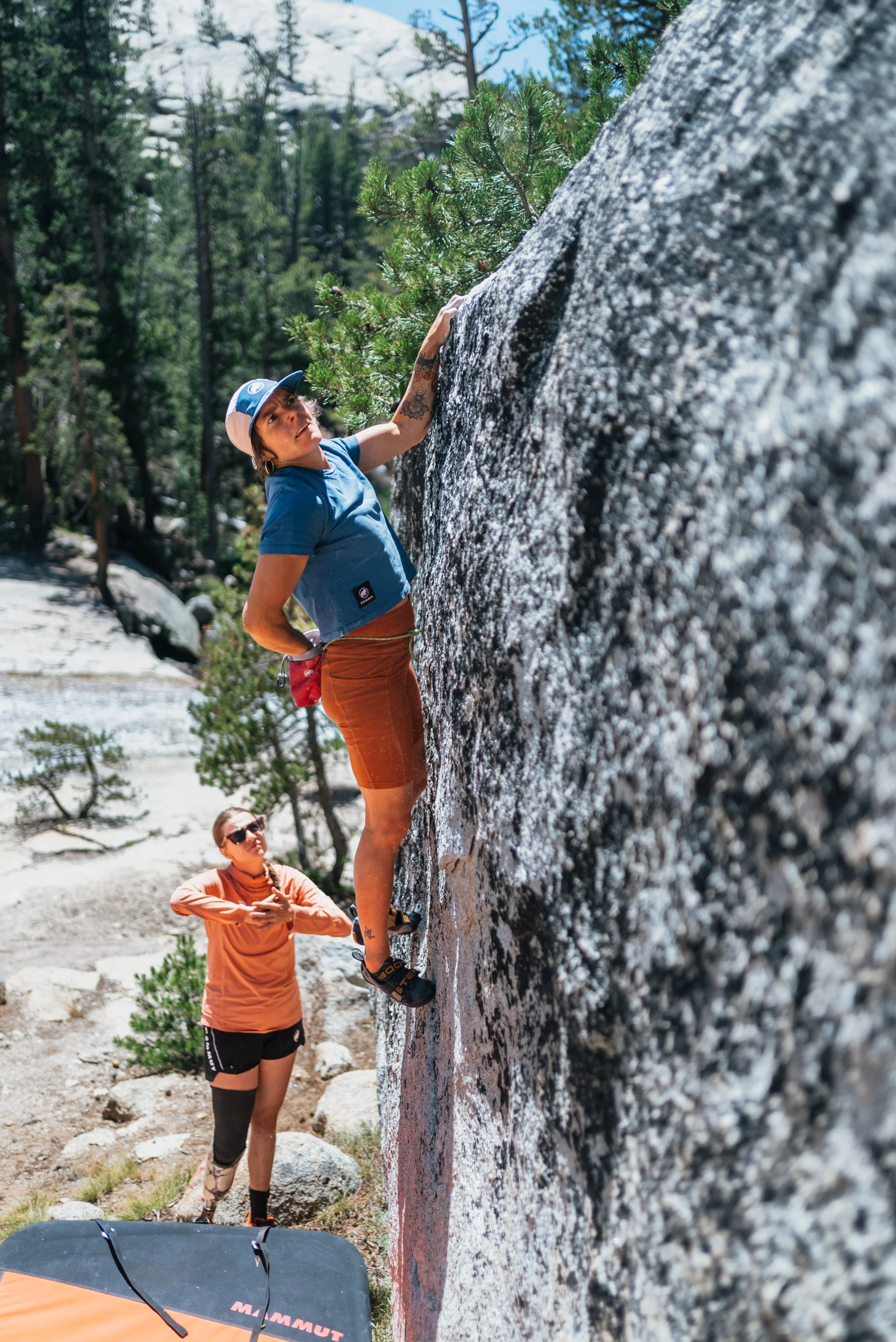A woman rock climbing on a large granite boulder outdoors with another woman spotting her from below. The woman climbing is wearing a blue shirt, shorts, and a cap. The spotter is wearing a pink top, black shorts, and sunglasses, standing on rocky gr