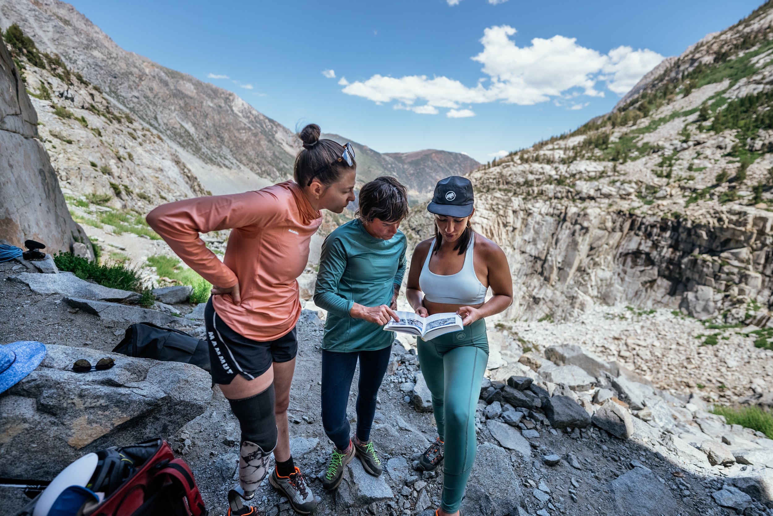 Three women hikers looking at an outdoor map in a mountainous area with rocky terrain and a partly cloudy sky.