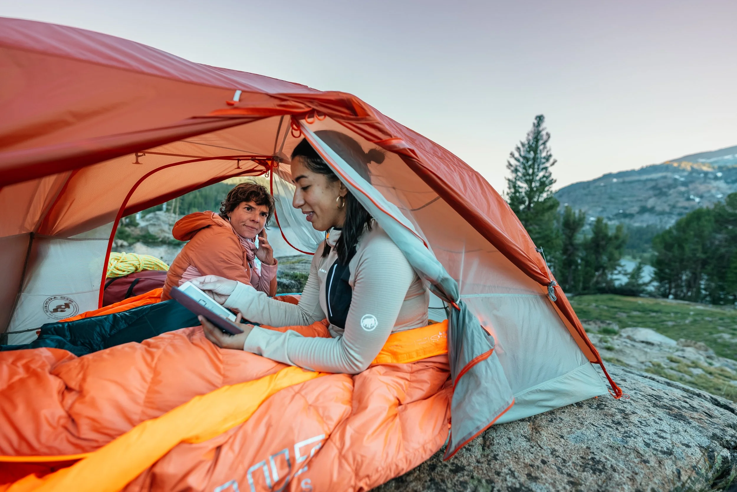 Two women inside an orange and gray camping tent in a mountainous outdoor setting, one is reading a book, and the other is looking at her, with trees and mountains in the background.