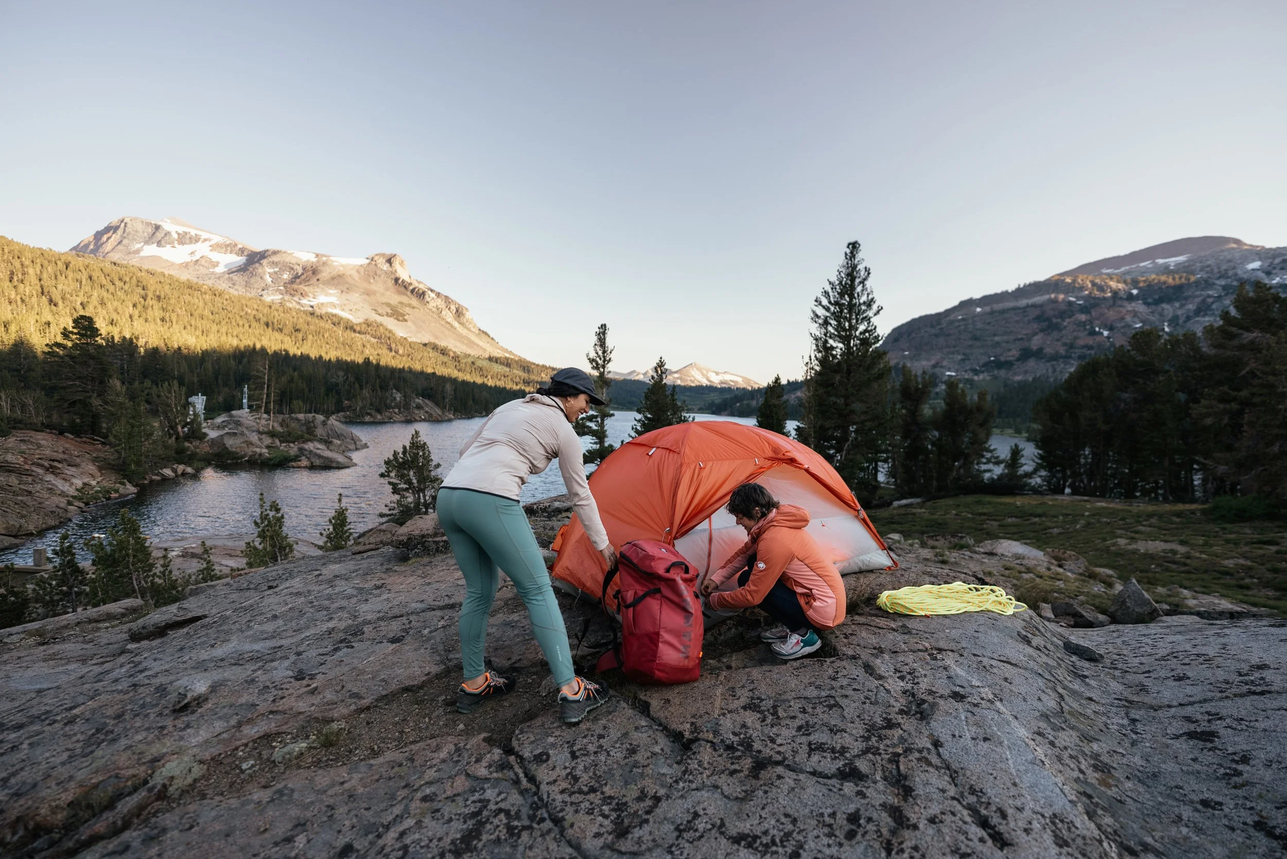 Two women setting up a tent on a rocky surface near a lake with mountains and pine trees in the background.