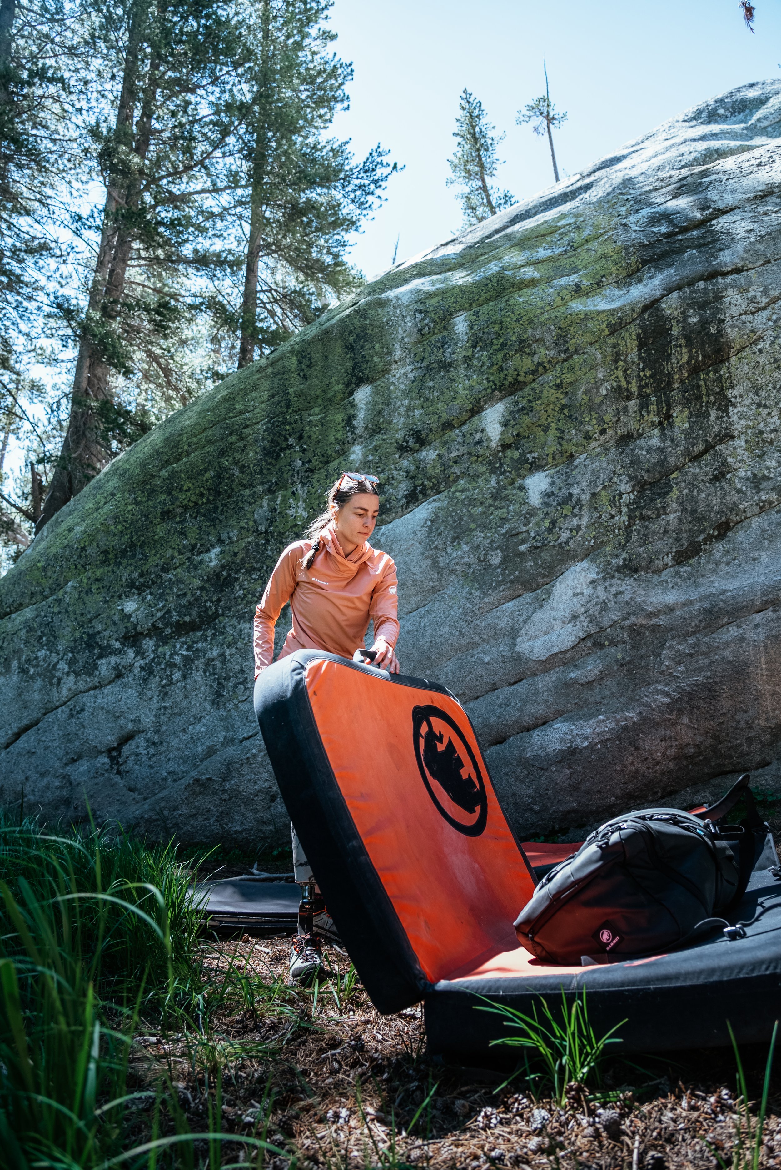 A woman in outdoor gear standing in a forested area next to a large mossy rock, preparing equipment.