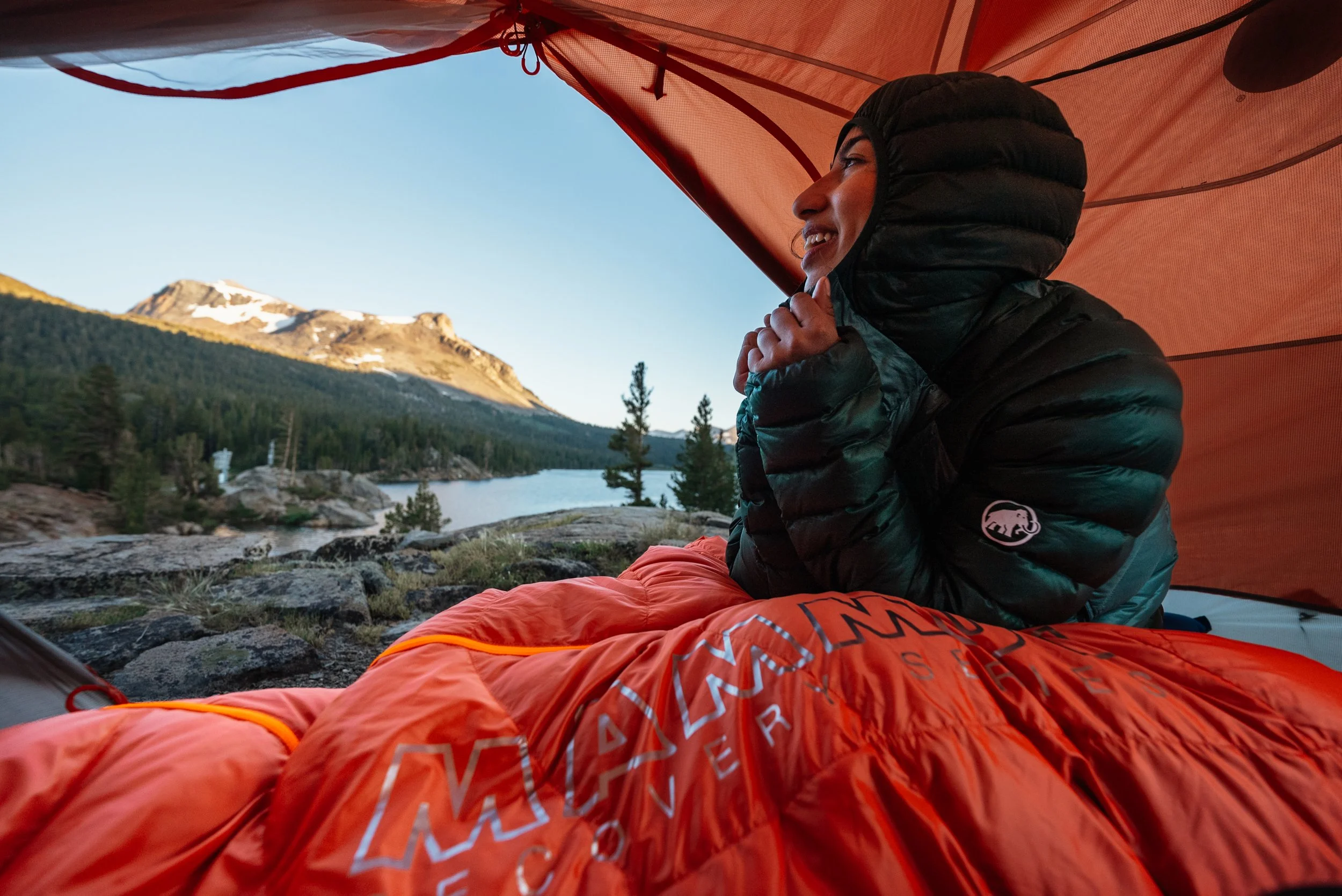 A person sitting inside a tent near a lake with mountains and trees in the background, dressed in outdoor camping gear, smiling and looking to the side.
