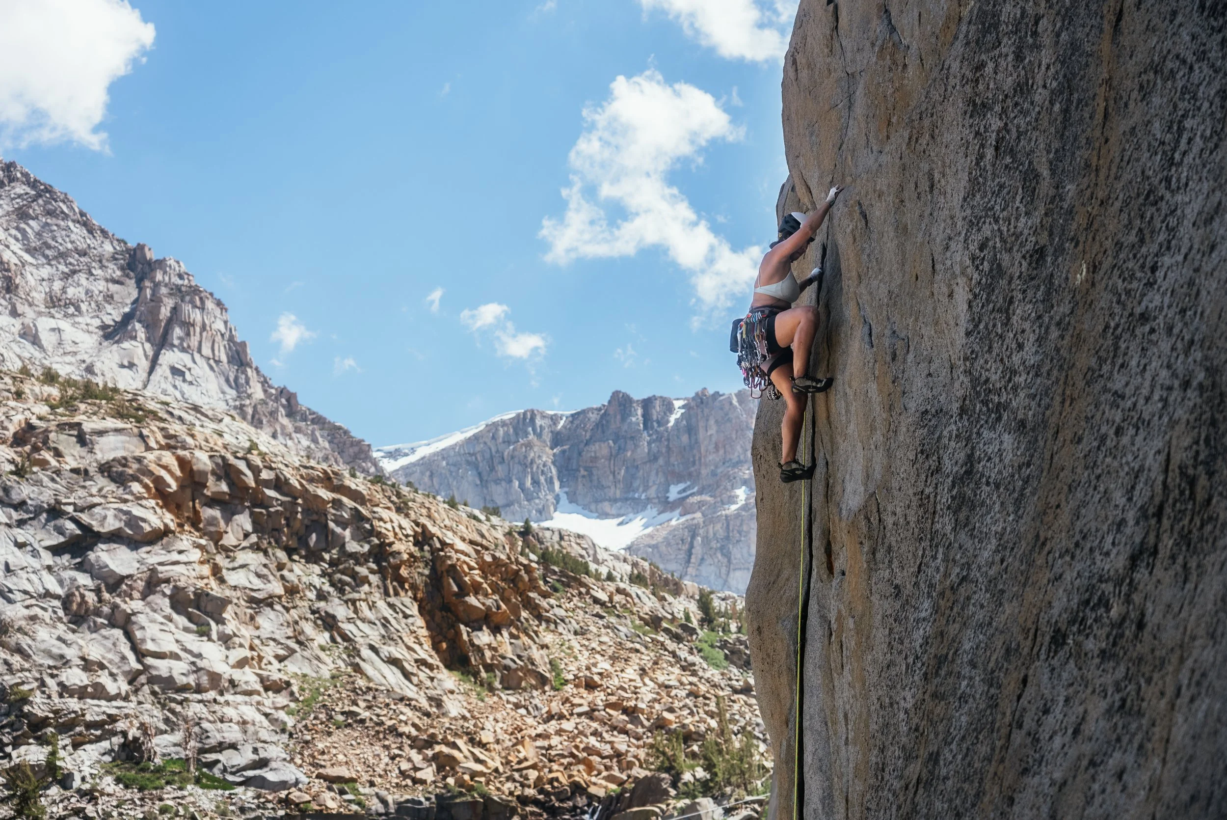 A rock climber ascending a tall vertical rock face in a mountainous area with snow-capped peaks in the background under a blue sky with some clouds.
