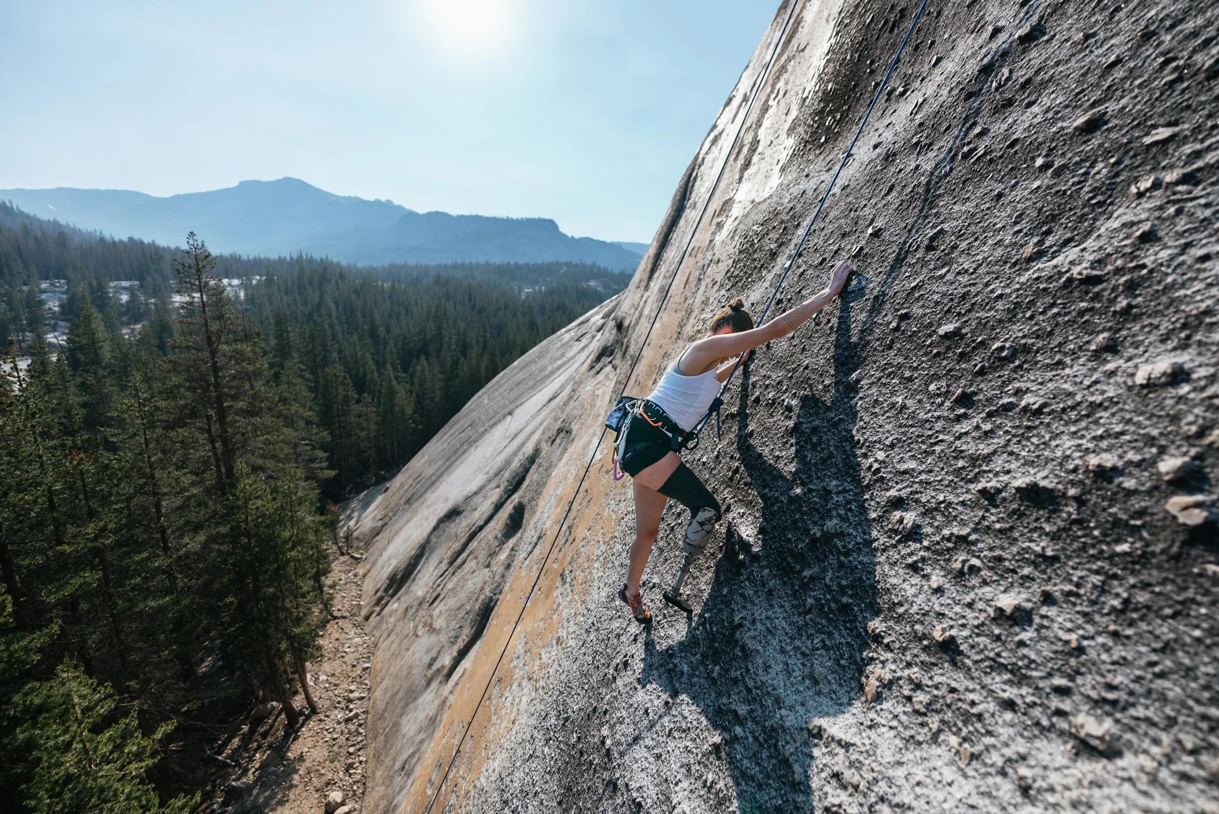 A woman with a prosthetic leg rock climbing a steep granite surface outdoors during daytime, with a forest and mountain range in the background.