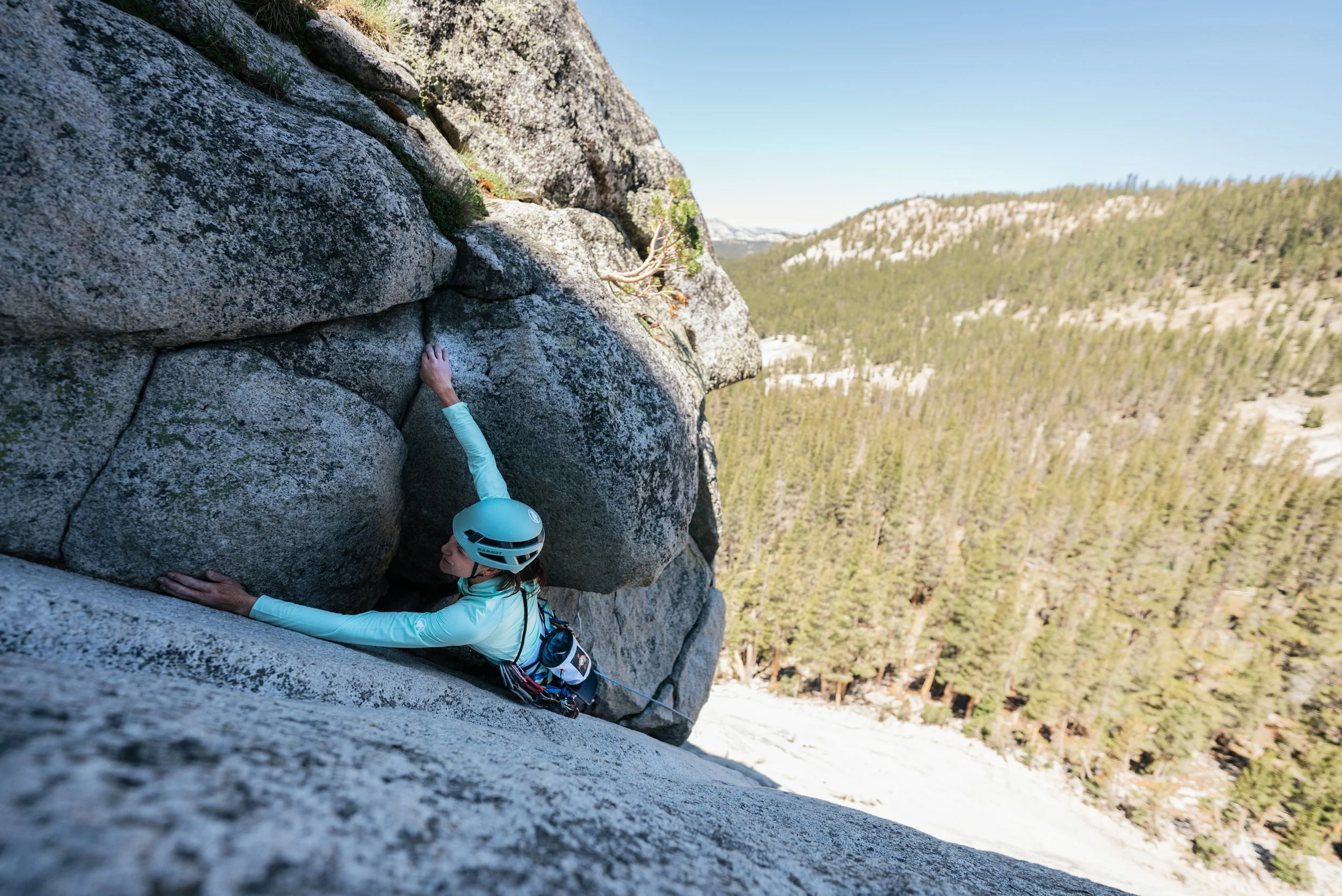A woman wearing a helmet and outdoor apparel rock climbing on a steep granite wall in a mountainous forested area.