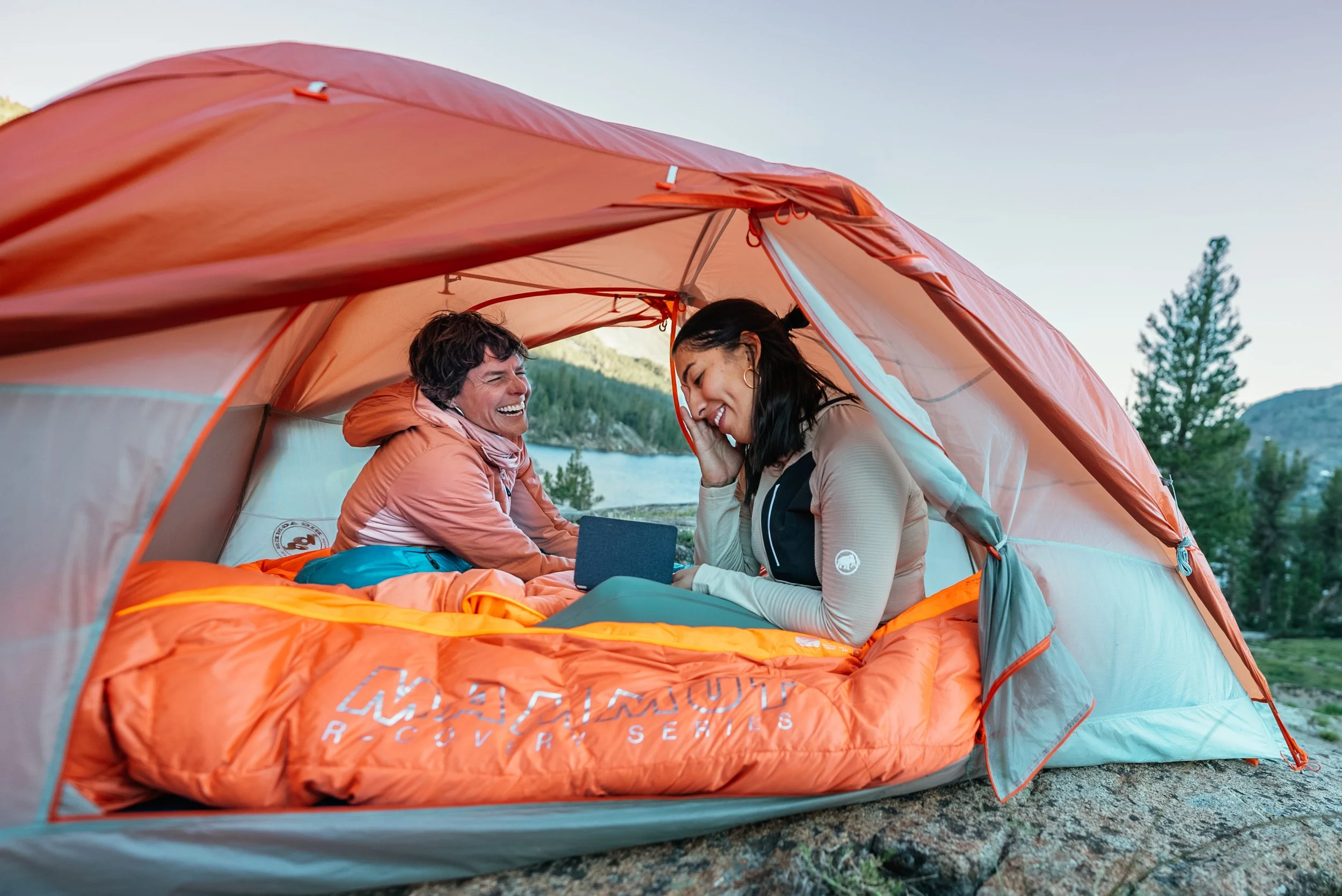 Two women inside an orange and gray tent, smiling and talking with a scenic lake and forest in the background during daytime.