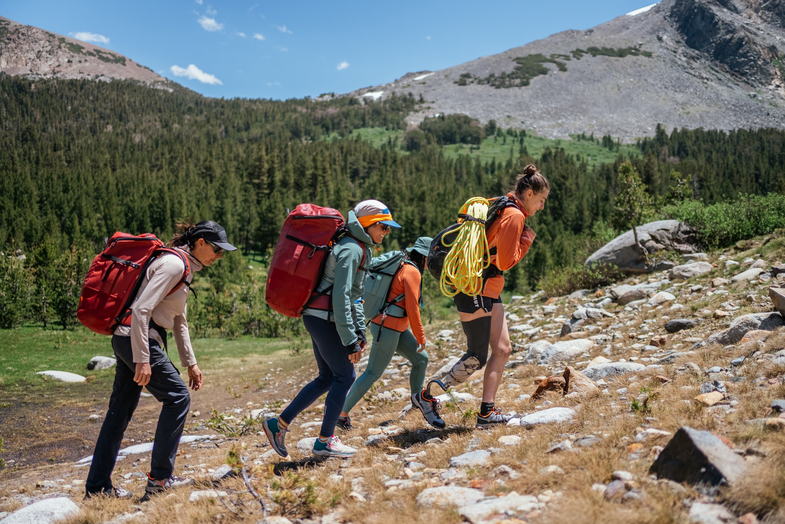 Four women hiking uphill on rocky terrain in a mountainous area with green trees and a blue sky.