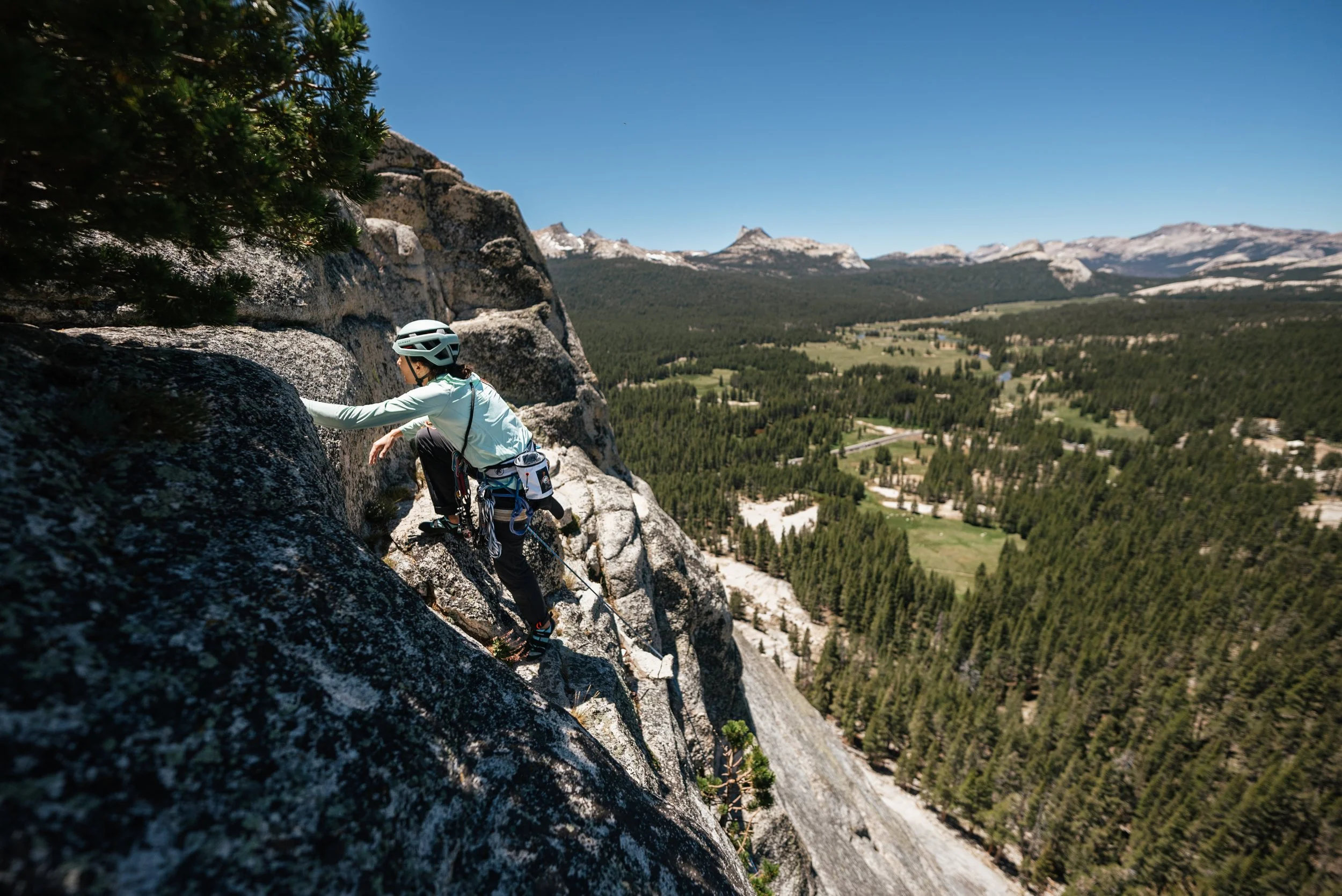 A person wearing a helmet and climbing gear scaling a steep rocky cliff, overlooking a lush green valley with trees and distant mountains under a clear blue sky.