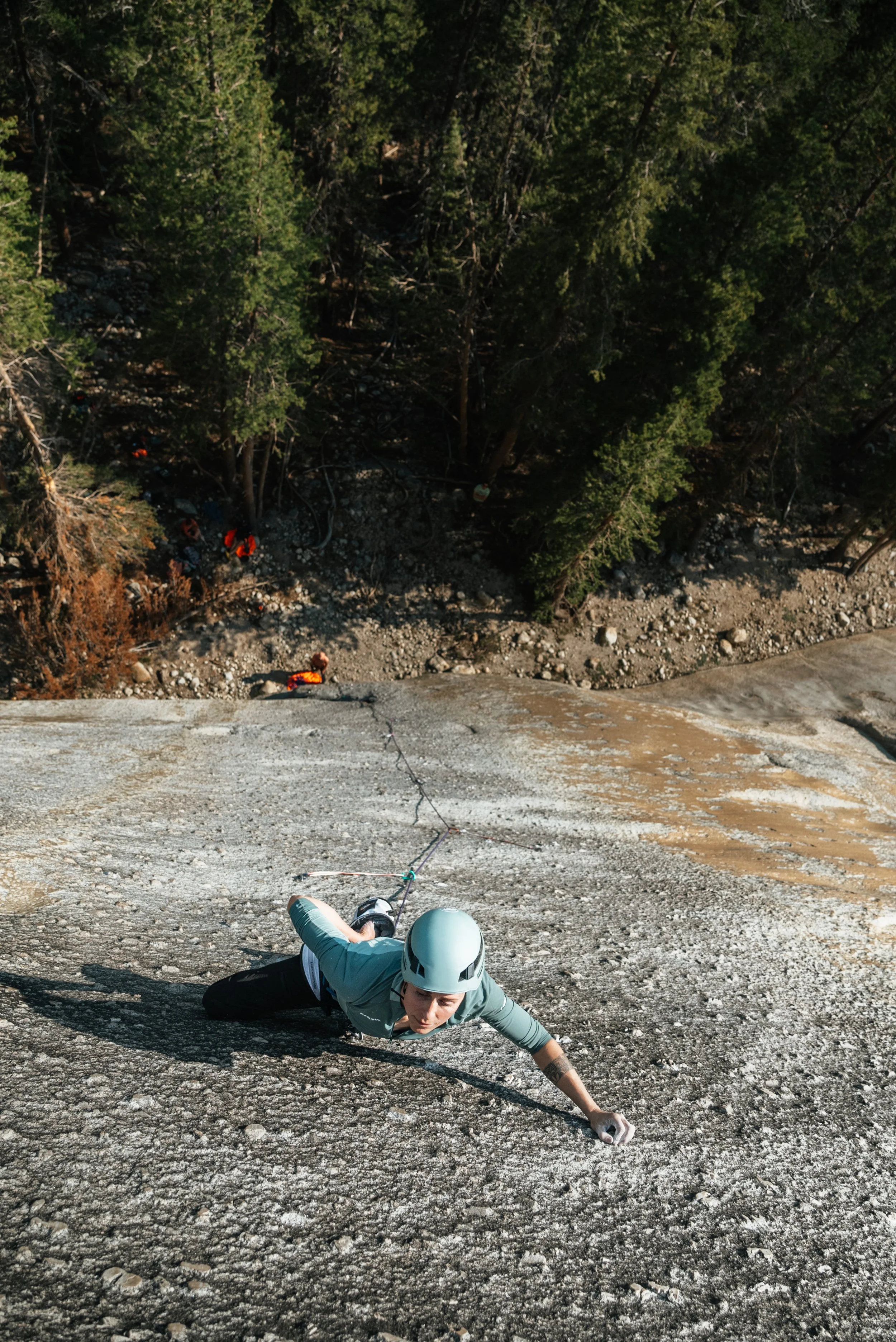 A person rock climbing on a steep granite wall wearing a helmet and climbing gear, with trees and rocky terrain below.