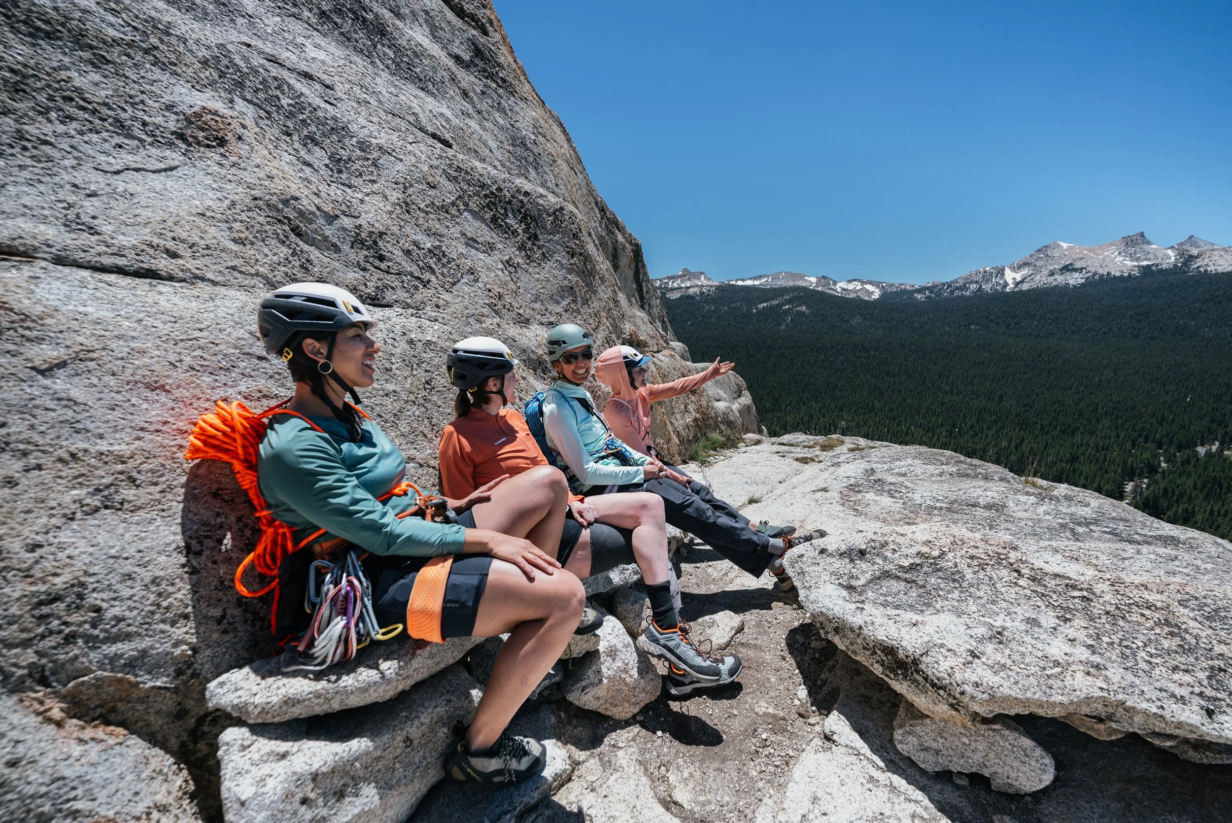 Four women in climbing gear and helmets sit on a large rock ledge, enjoying a view of a forested mountain landscape and clear blue sky.