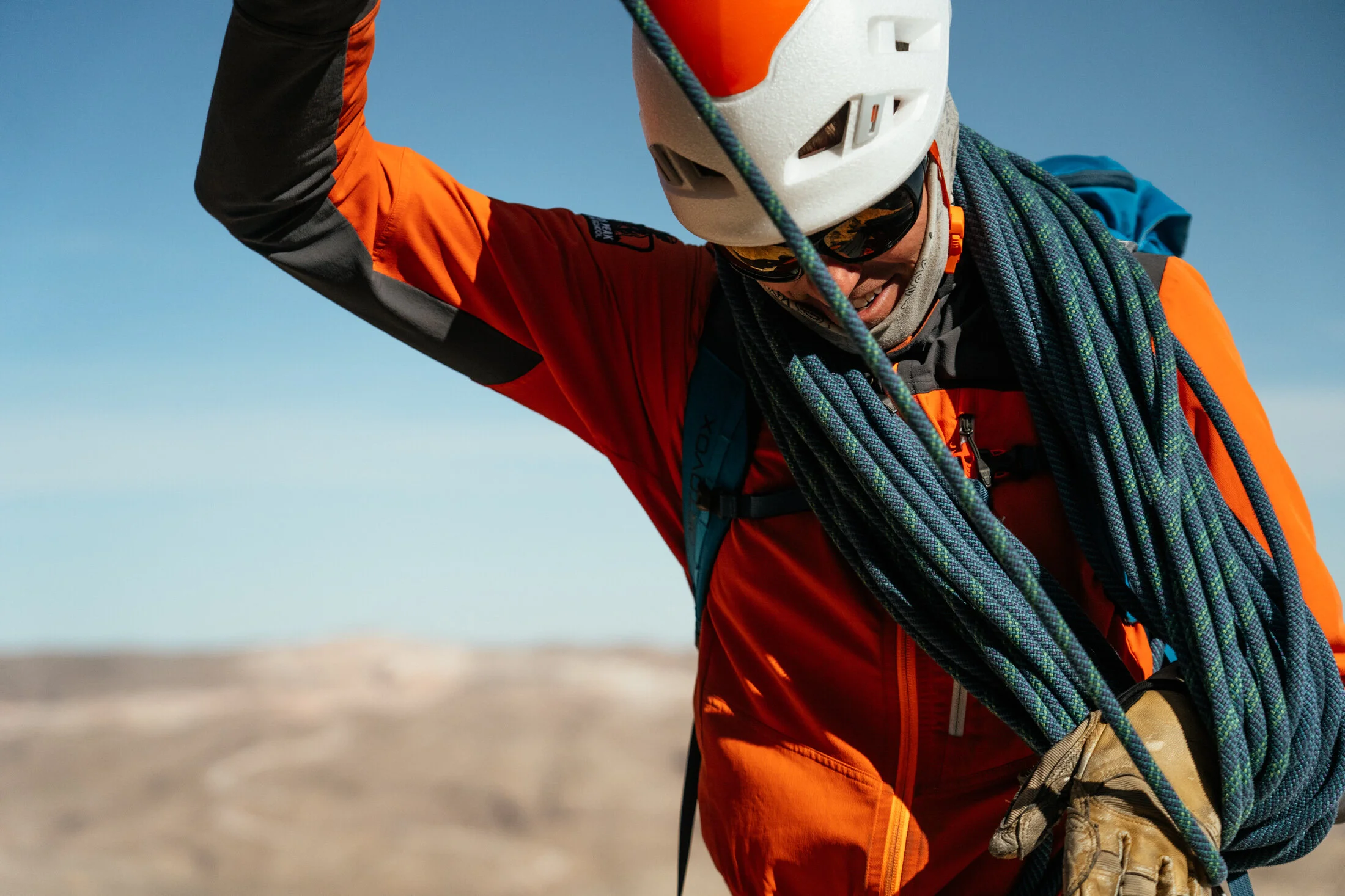 A rock climber wearing a white helmet, orange and black jacket, and sunglasses, carrying climbing ropes on his shoulder.
