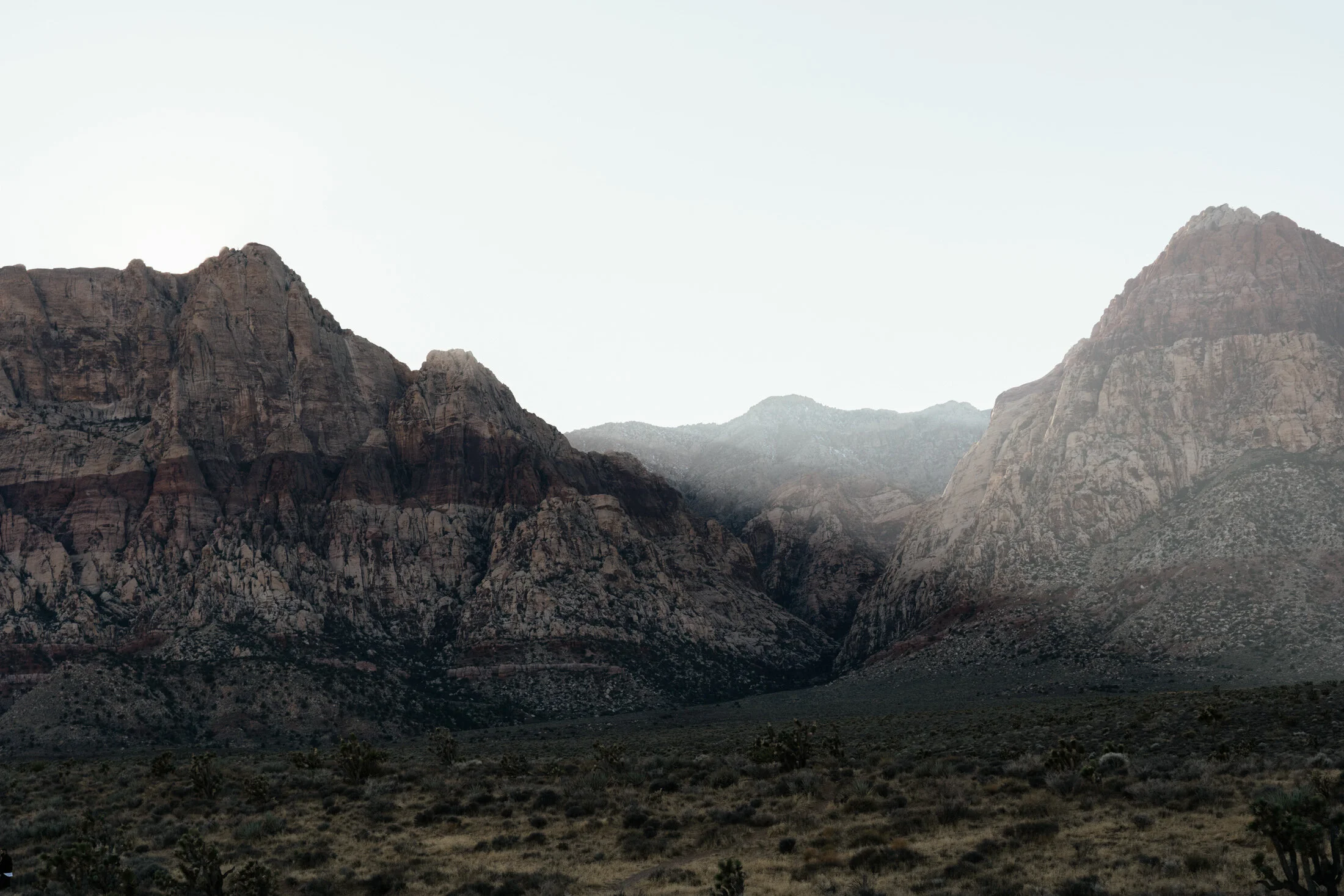 A rugged mountain landscape with multiple large rock formations and sparse desert vegetation in the foreground.