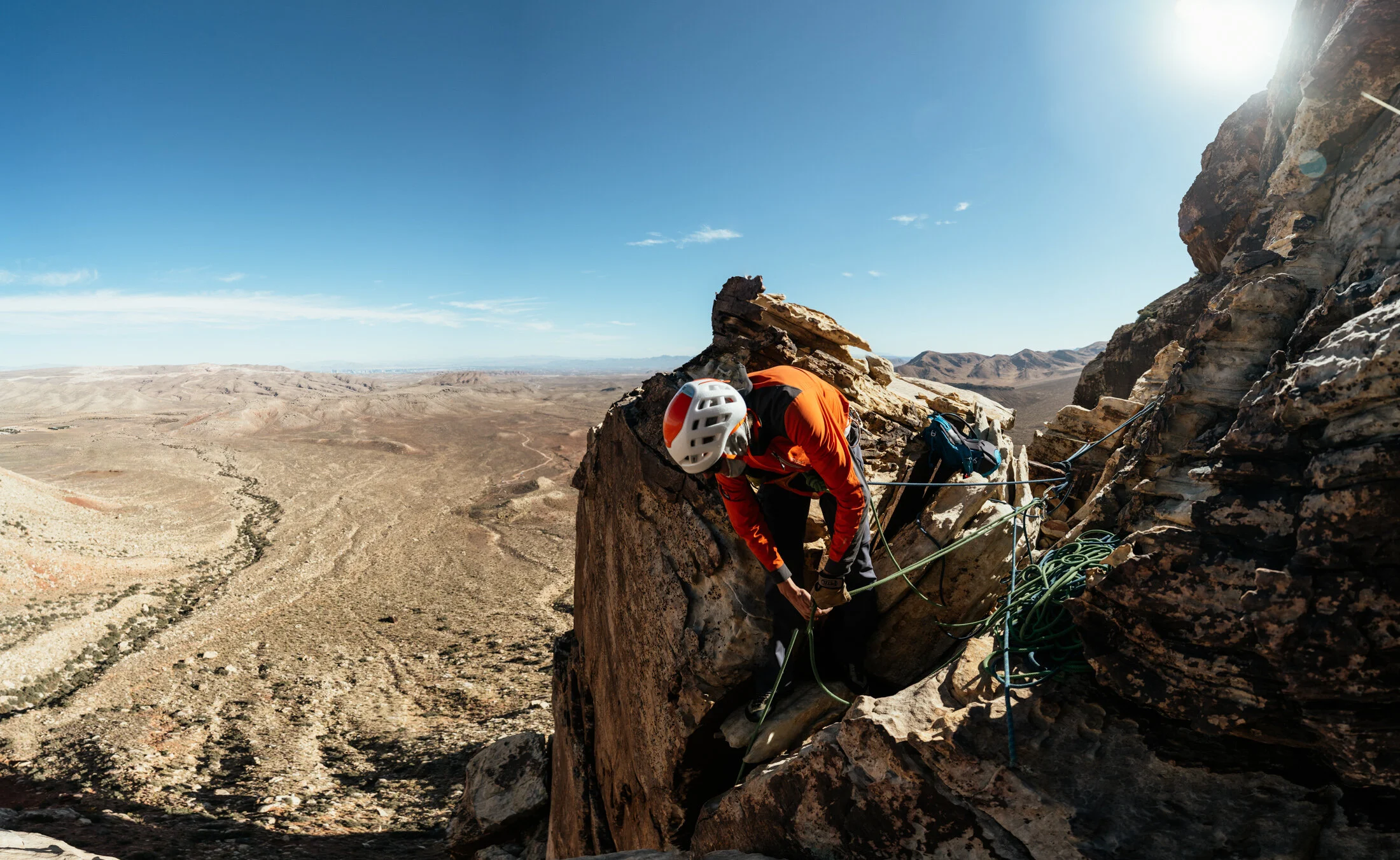 A person wearing a white helmet and orange jacket is climbing a steep rock face with safety ropes in a desert landscape under a clear blue sky.