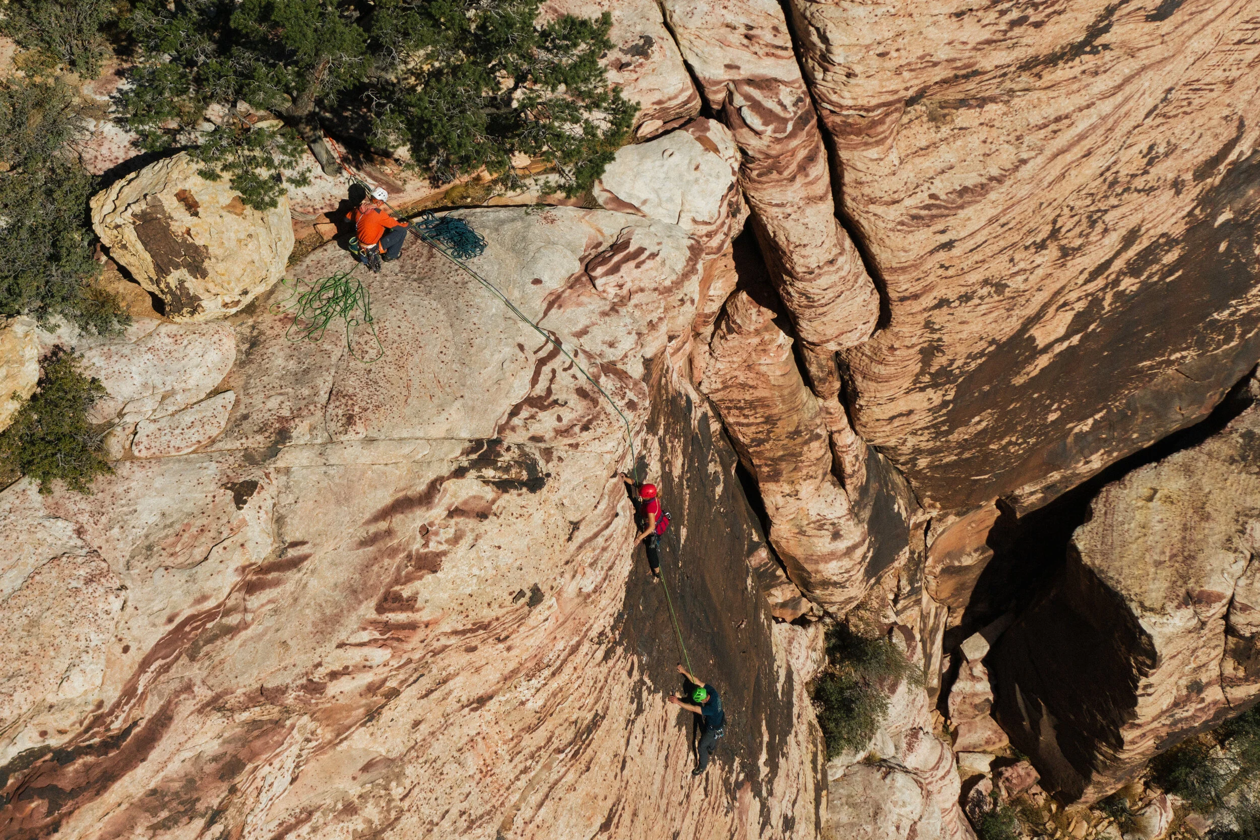 Two rock climbers scaling a steep, layered sandstone cliff with assistance from a rope, while a third person manages the rope at the top of the cliff.