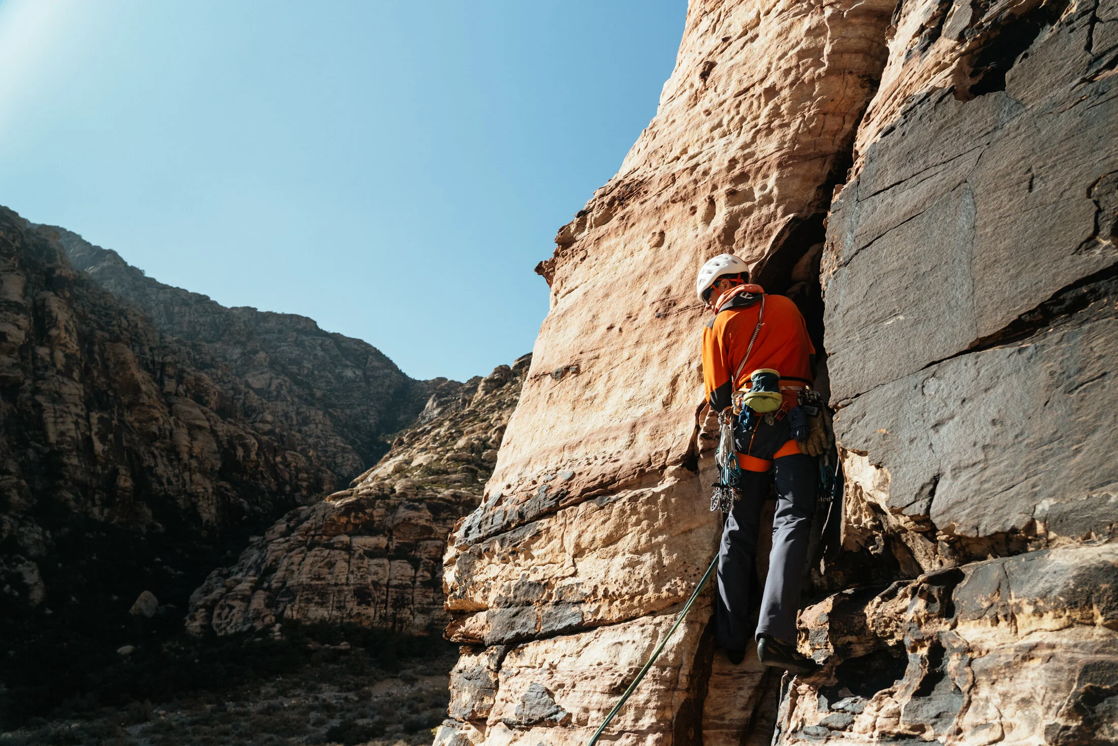 A person wearing a white helmet and orange jacket rock climbing on a rugged cliff face in a mountainous area under a clear blue sky.