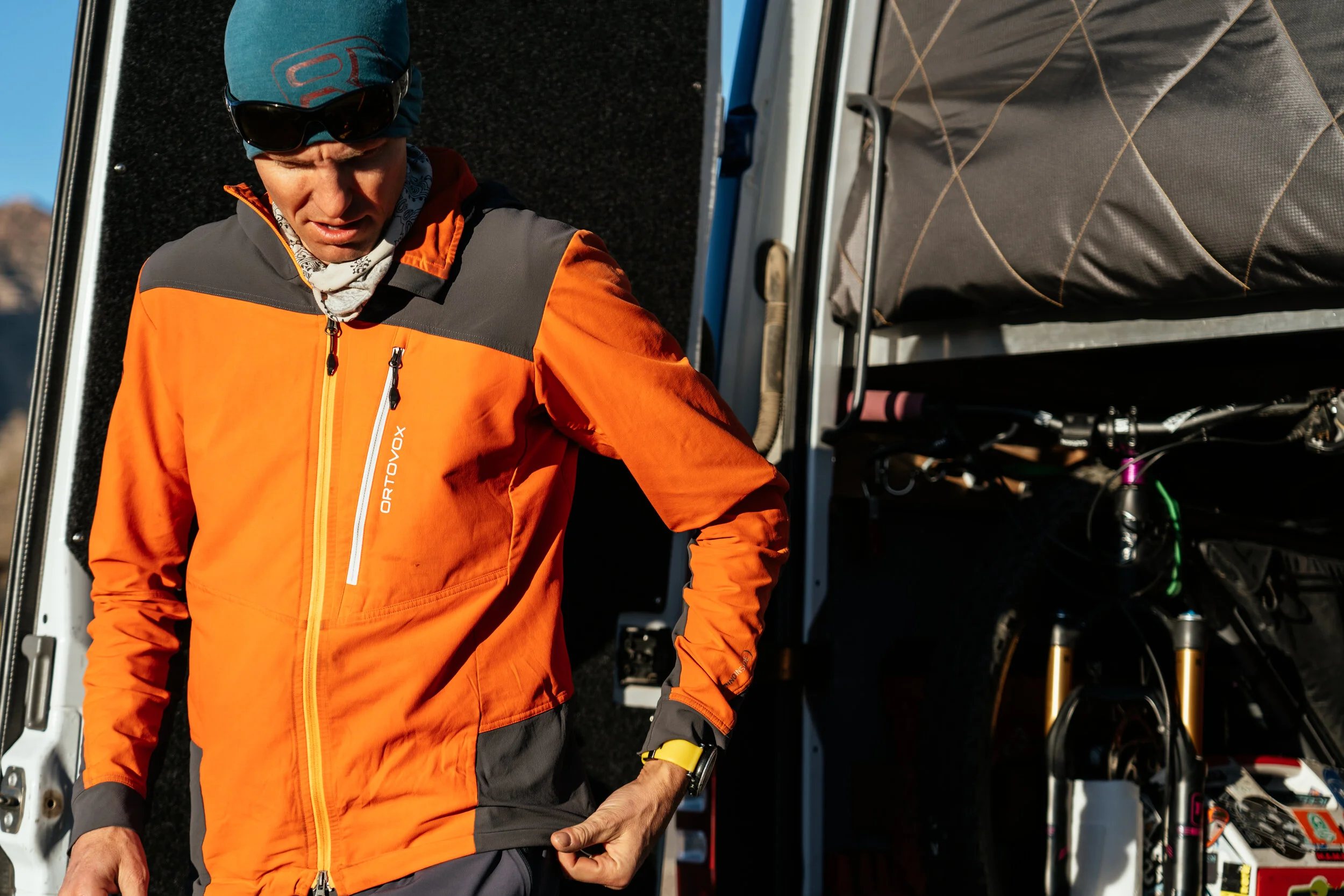 A man wearing an orange and gray jacket, a teal headband, sunglasses, and a white neck gaiter, standing near a mountain bike and van accessories.