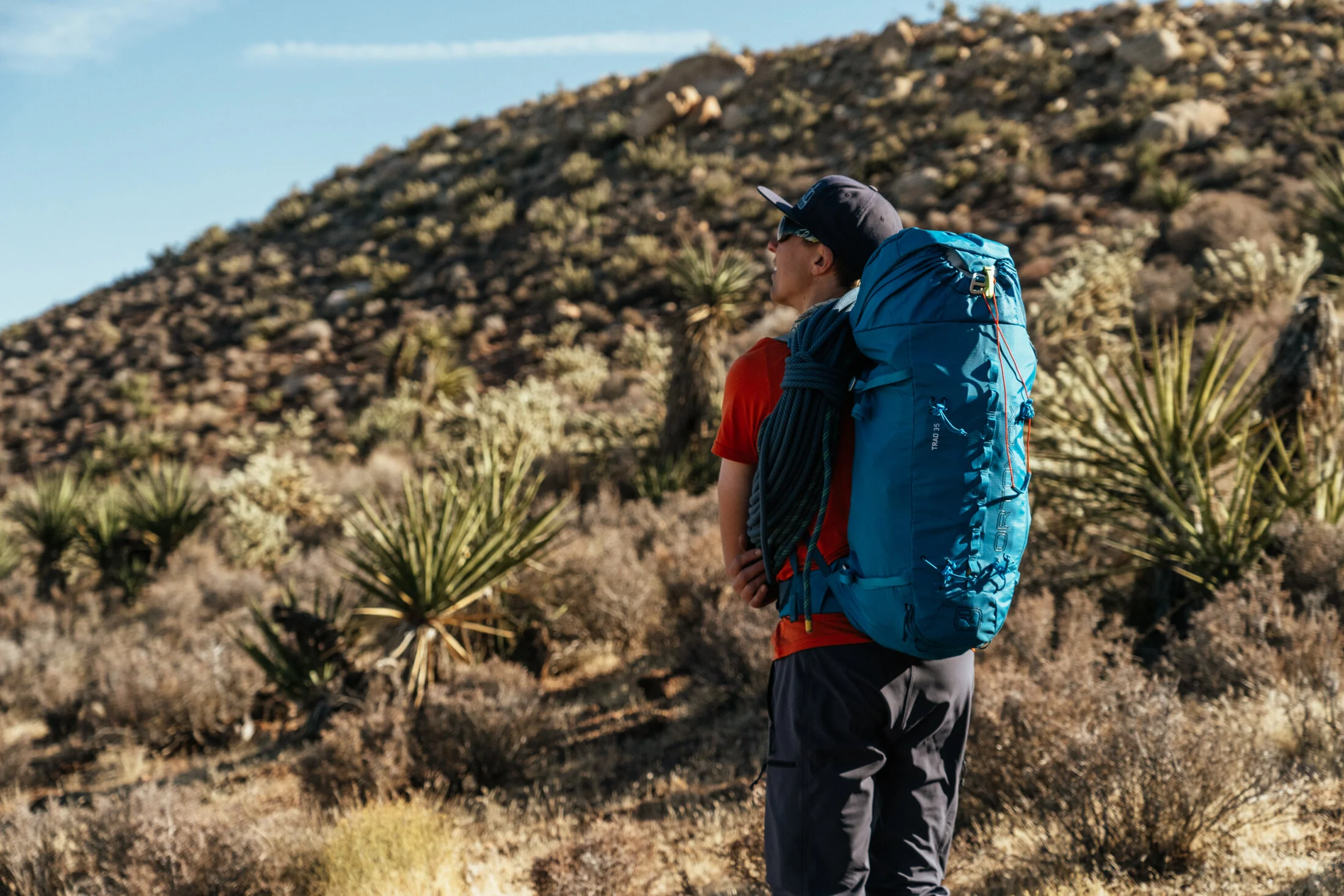 A man with a backpack hiking in a desert landscape with sparse vegetation and a hill in the background.