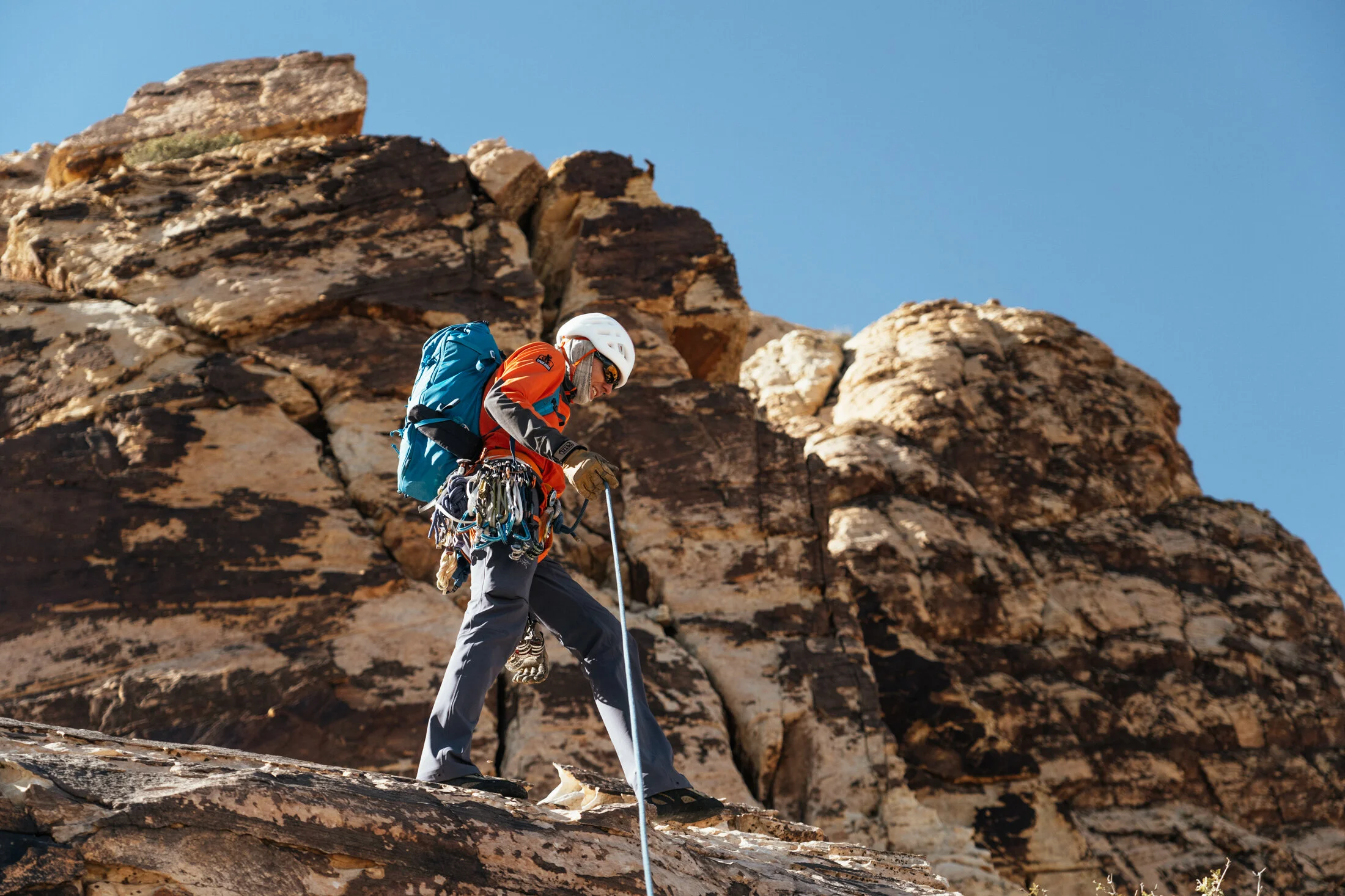 A male mountaineer climbing a rocky mountain slope, wearing a helmet, sunglasses, and carrying a large blue backpack.