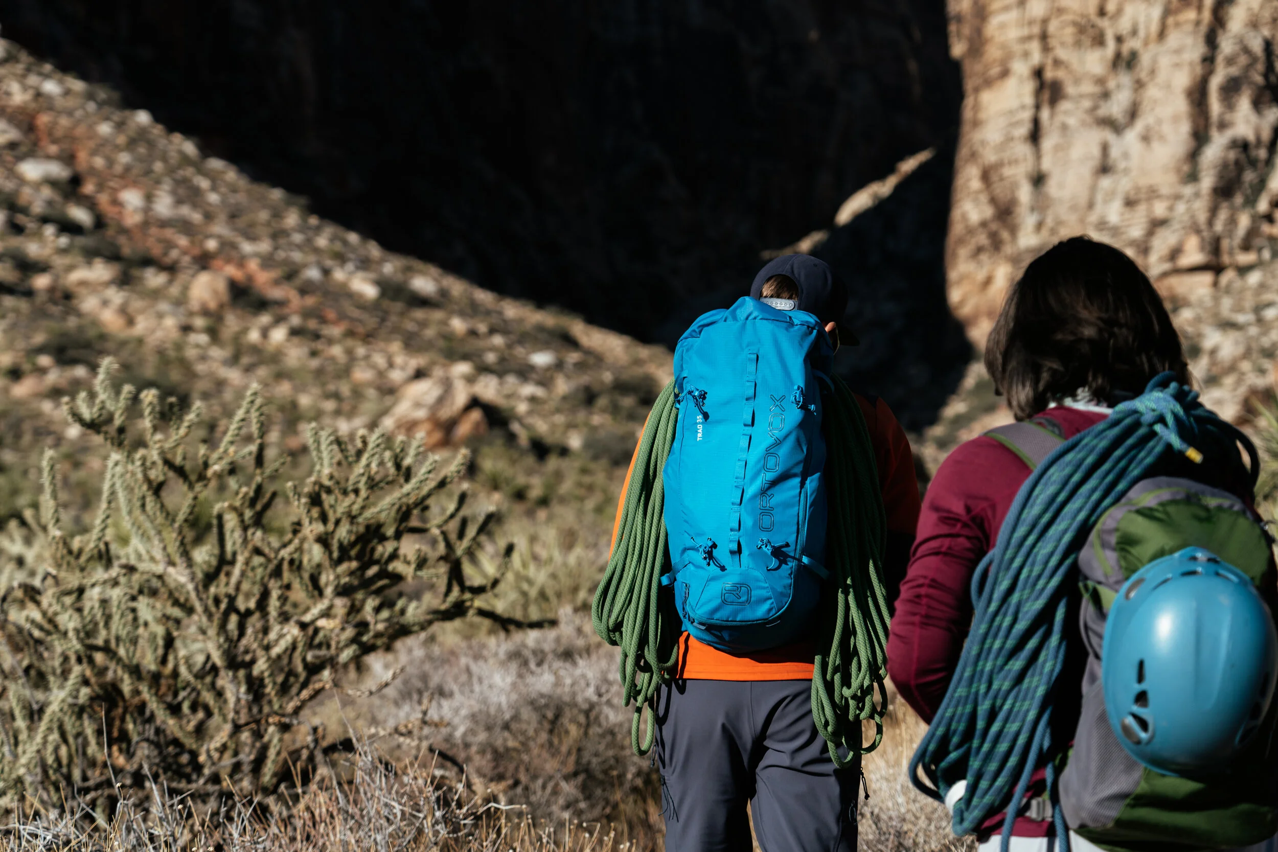 Two hikers walking through a desert landscape with cacti and rocky formations in the background, carrying backpacks and climbing gear.