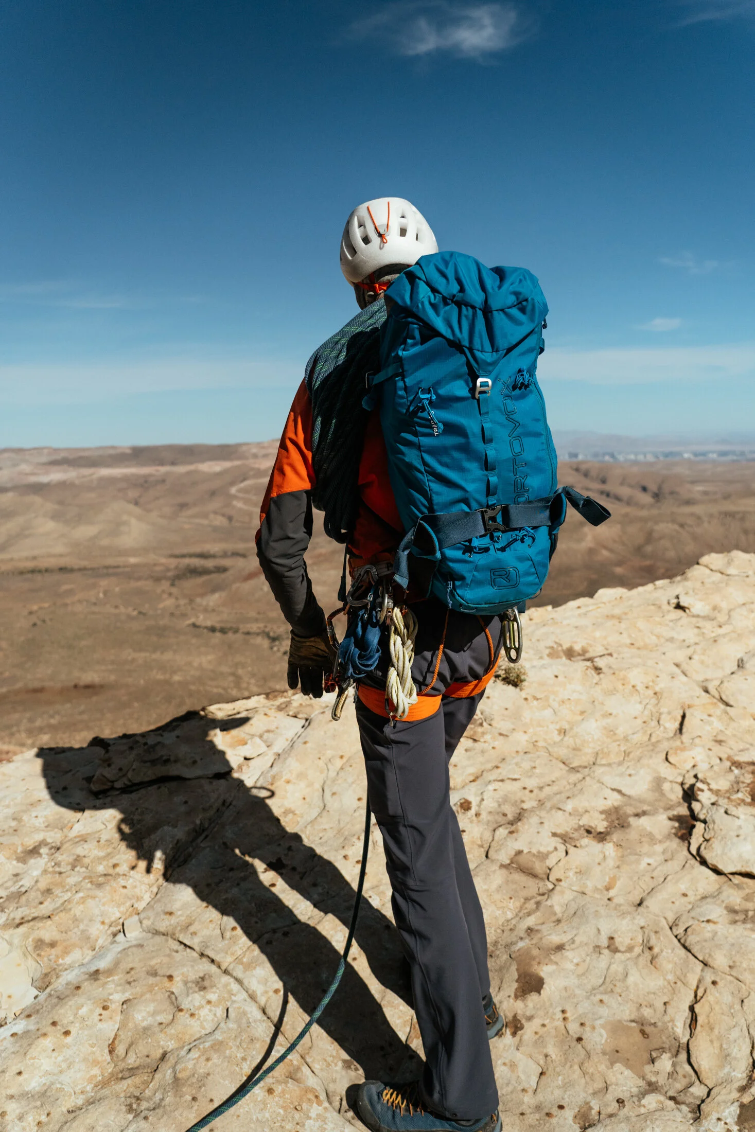 Back view of a climber on a rocky cliff, wearing a helmet, a blue backpack, and outdoor gear, overlooking a vast desert landscape under a blue sky.